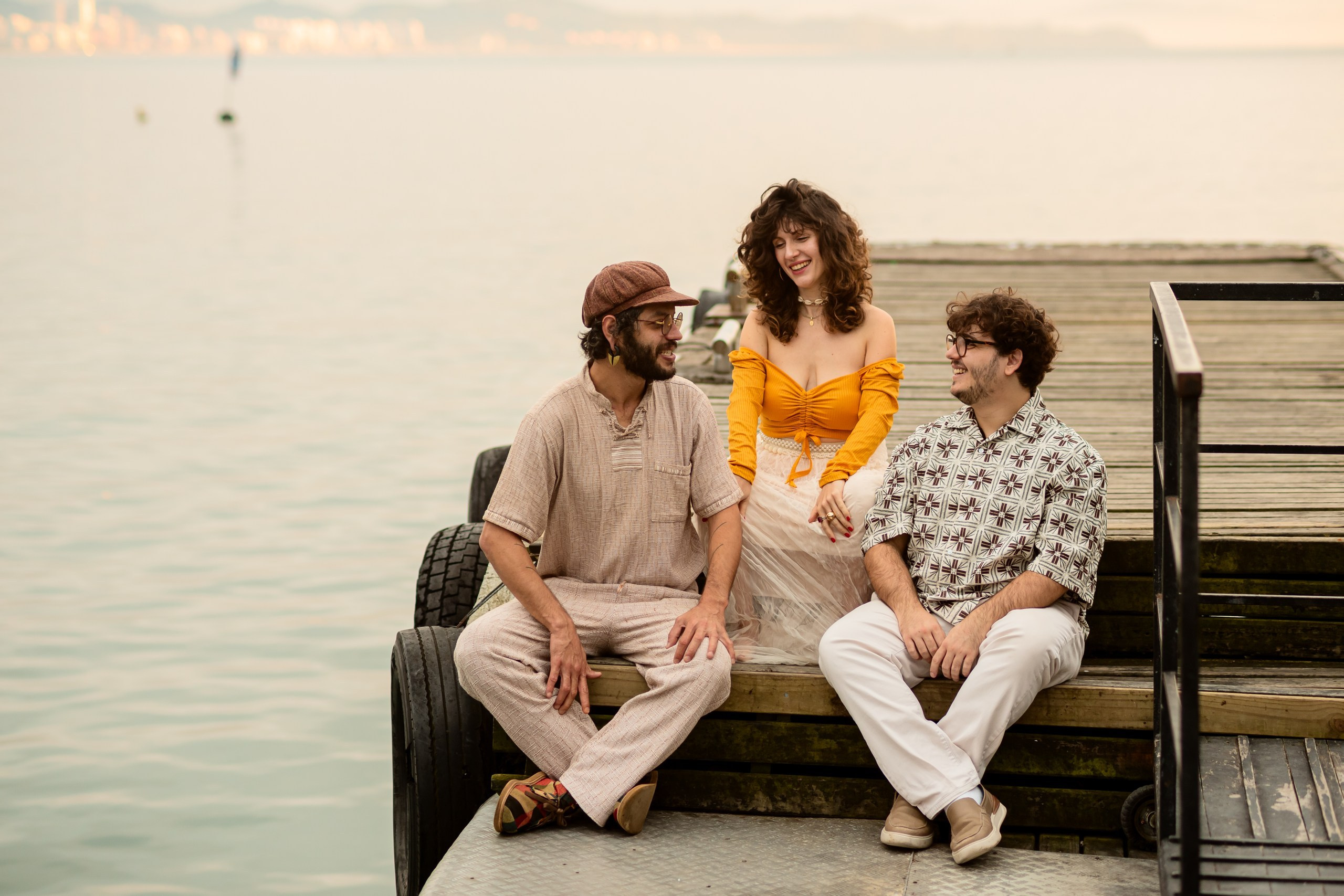 Ensaio fotográfico em Balneário Camboriú; cantora Joana Gaia e sua banda sentados no píer à beira da água na Praia de Laranjeiras para o álbum "Água Brasileira".