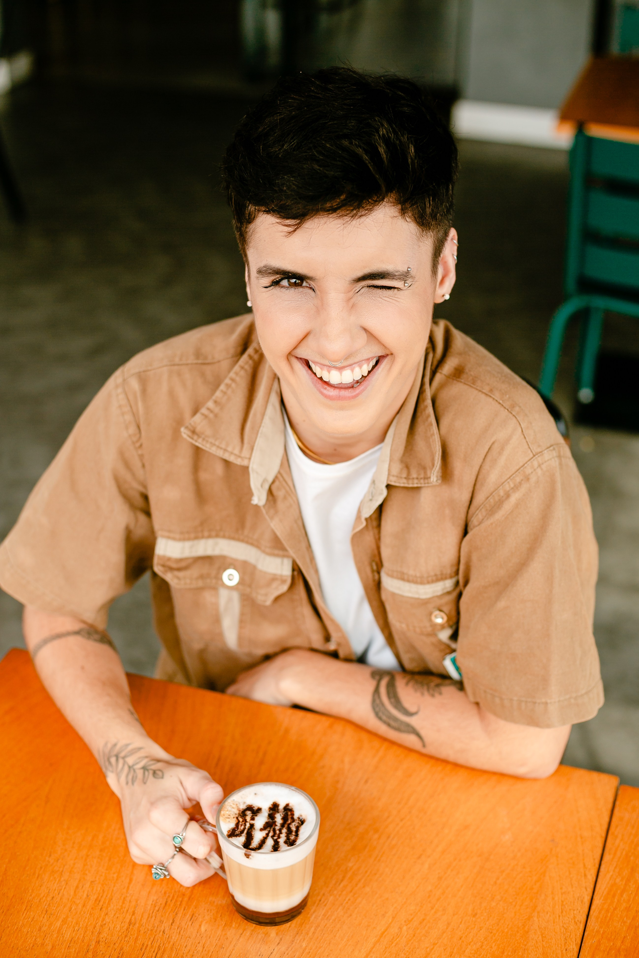 Fotógrafo de ensaio em Balneário Camboriú; retrato aconchegante e sorridente de jovem com cabelo curto escuro e camisa bege, segurando uma chávena de café decorada numa mesa de cafeteria.