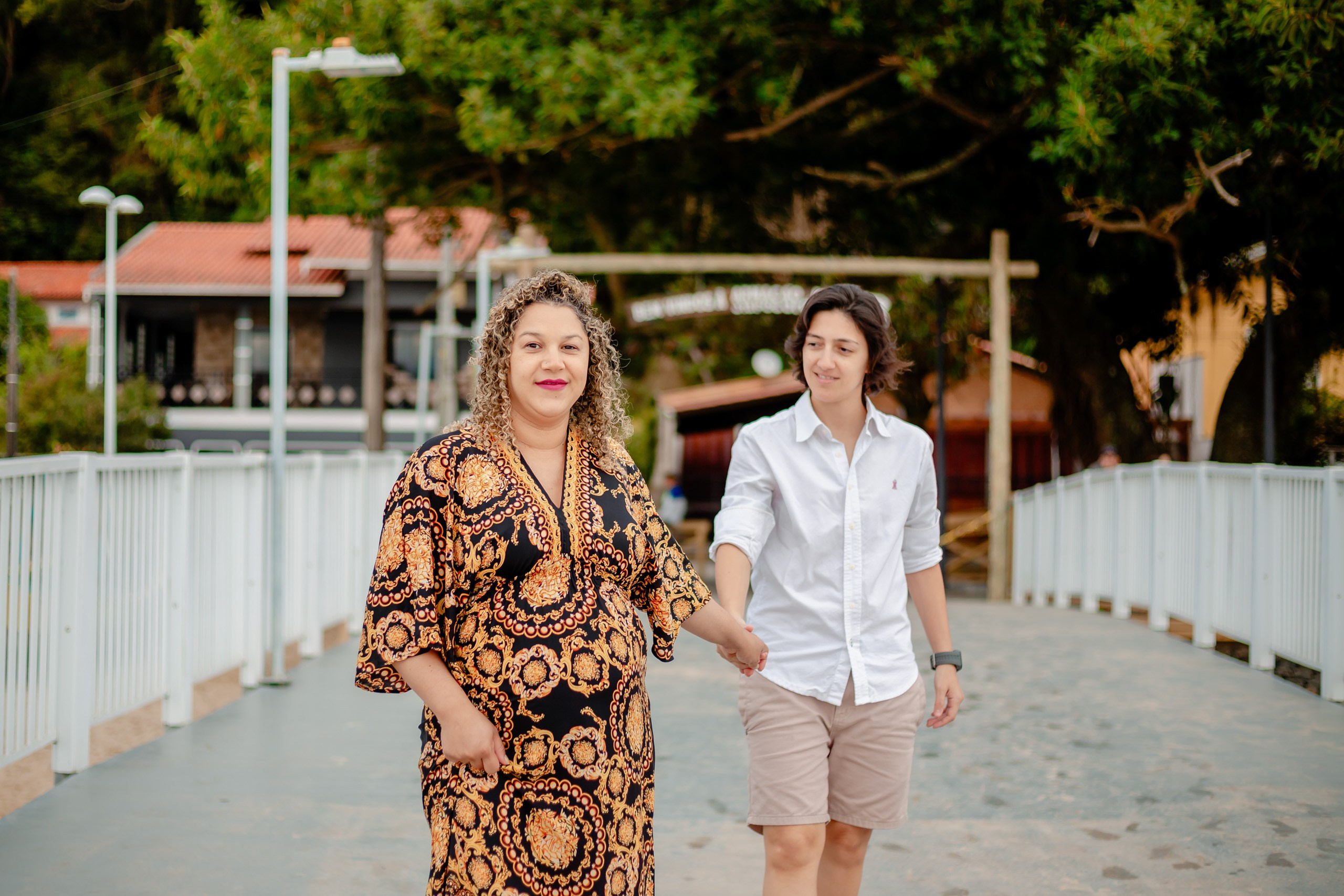 Fotógrafo ensaio gestante em Penha; casal caminhando de mãos dadas sorridentes em uma passarela ao ar livre, em ensaio lifestyle leve e afetuoso.