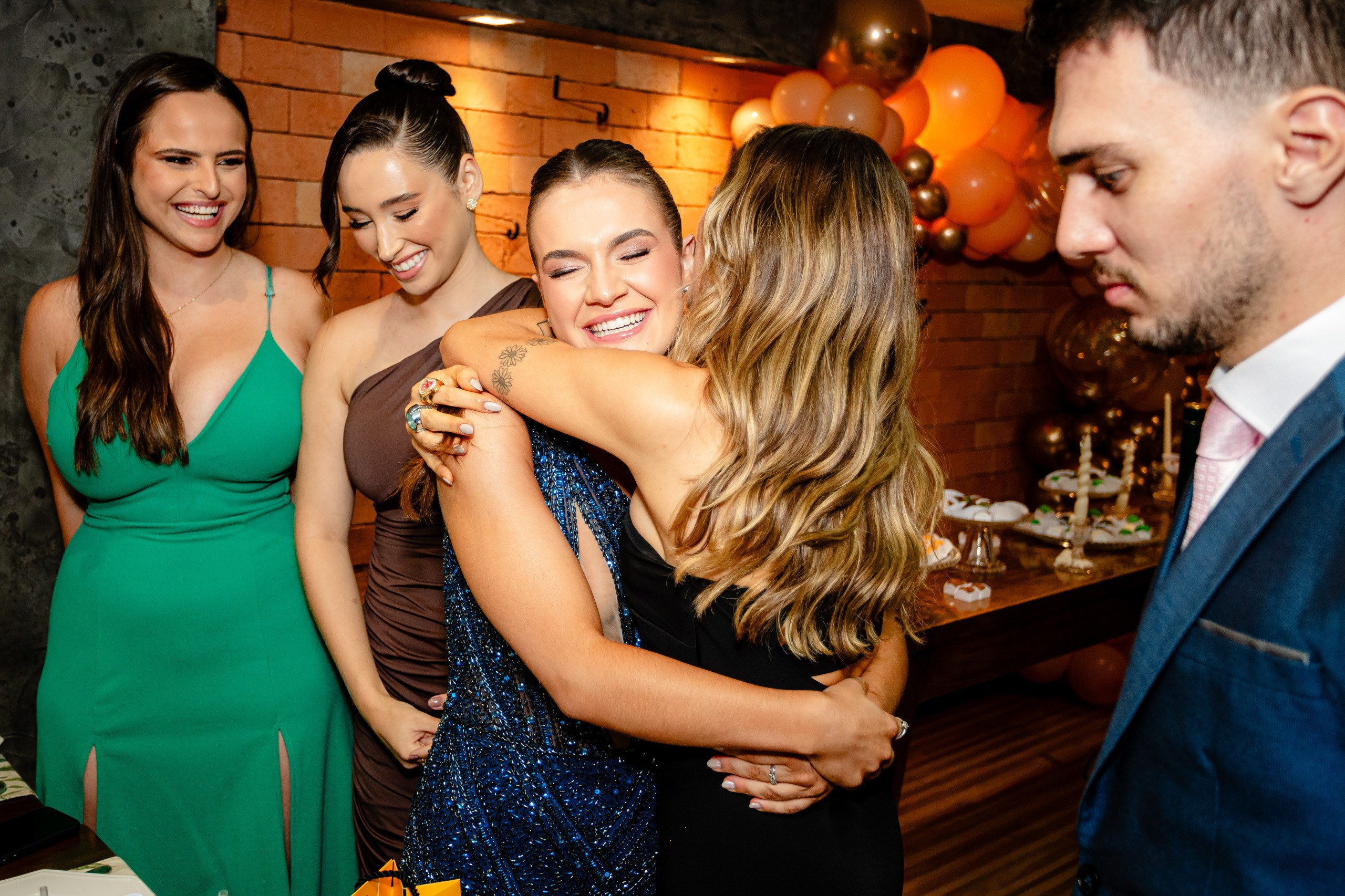 Fotógrafo de formatura em Balneário Camboriú; a formanda recebe um abraço caloroso de uma convidada, sorrindo ao lado de amigos durante a festa.