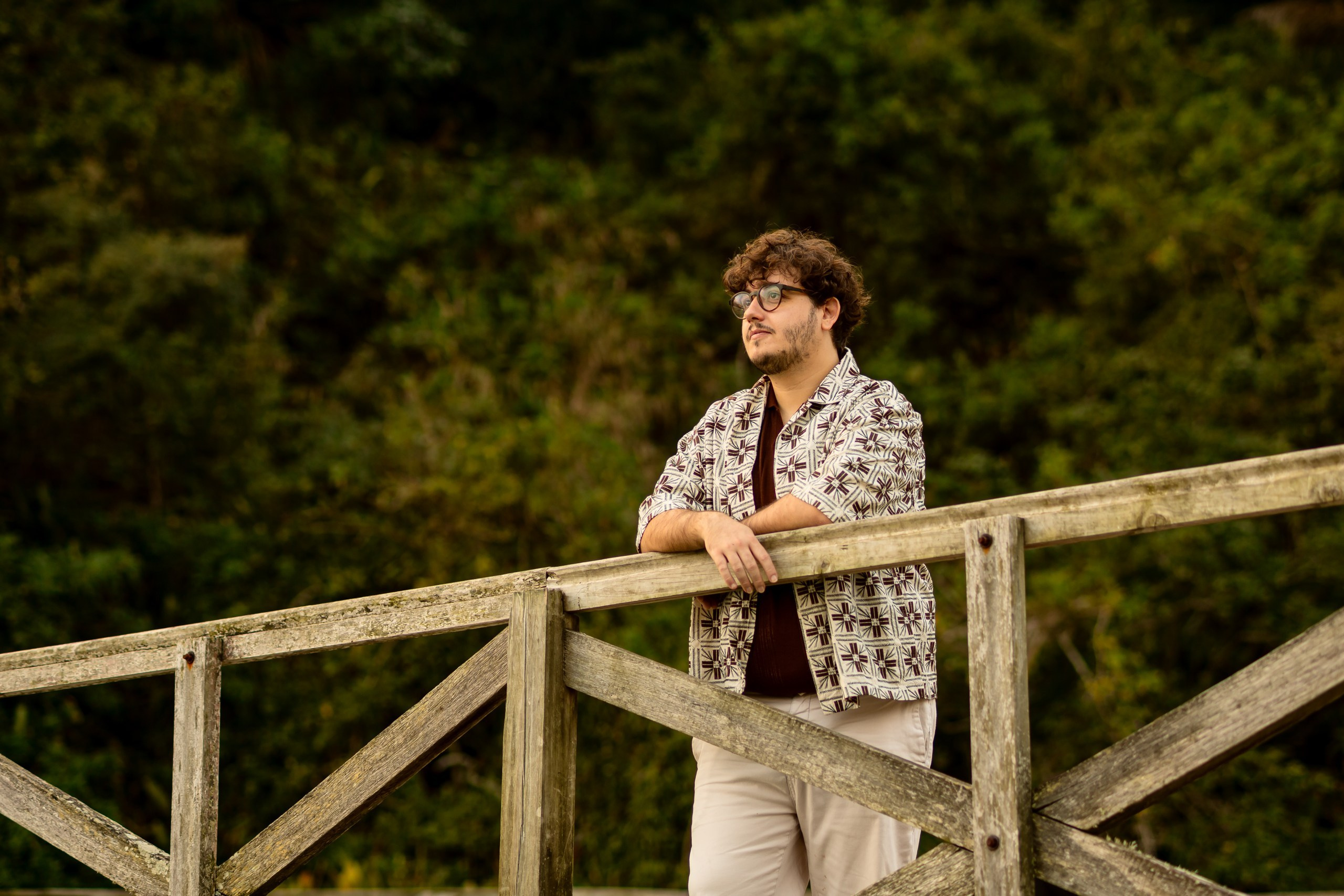 Ensaio fotográfico em Balneário Camboriú; retrato de integrante da banda de Joana Gaia apoiado no parapeito de madeira de uma ponte com vegetação verde ao fundo na Praia de Laranjeiras para o álbum "Água Brasileira".