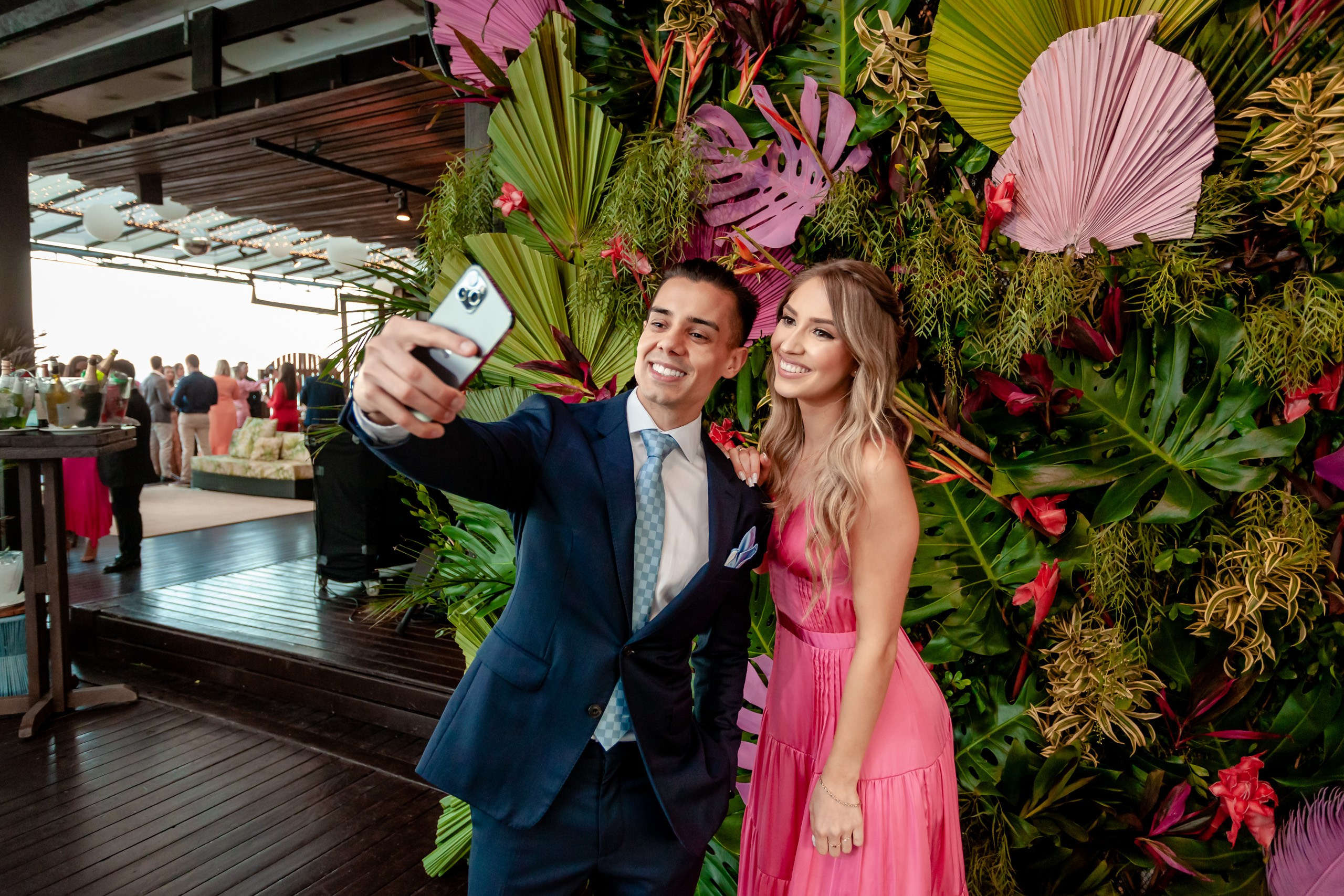 Fotógrafo festa de formatura em Balneário Camboriú; casal sorridente a tirar uma selfie divertida em frente ao painel de folhagens do Bali Bistro Beach.