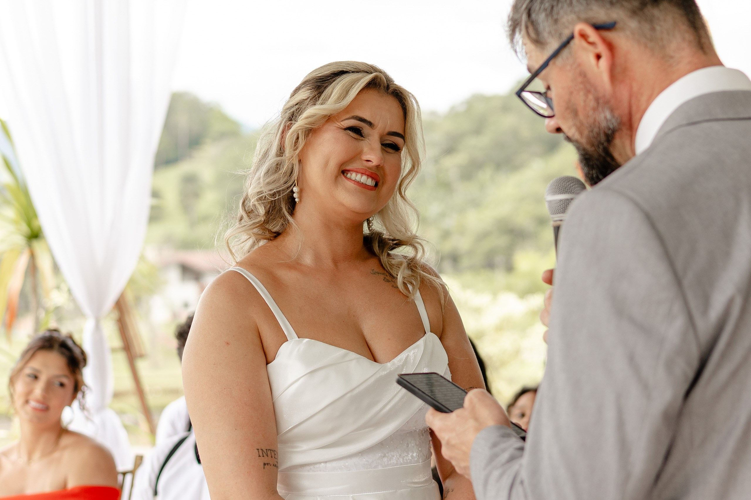 Fotógrafo de casamento em Santa Catarina; noiva sorridente olhando para o noivo enquanto ele lê os votos durante a cerimônia no campo.