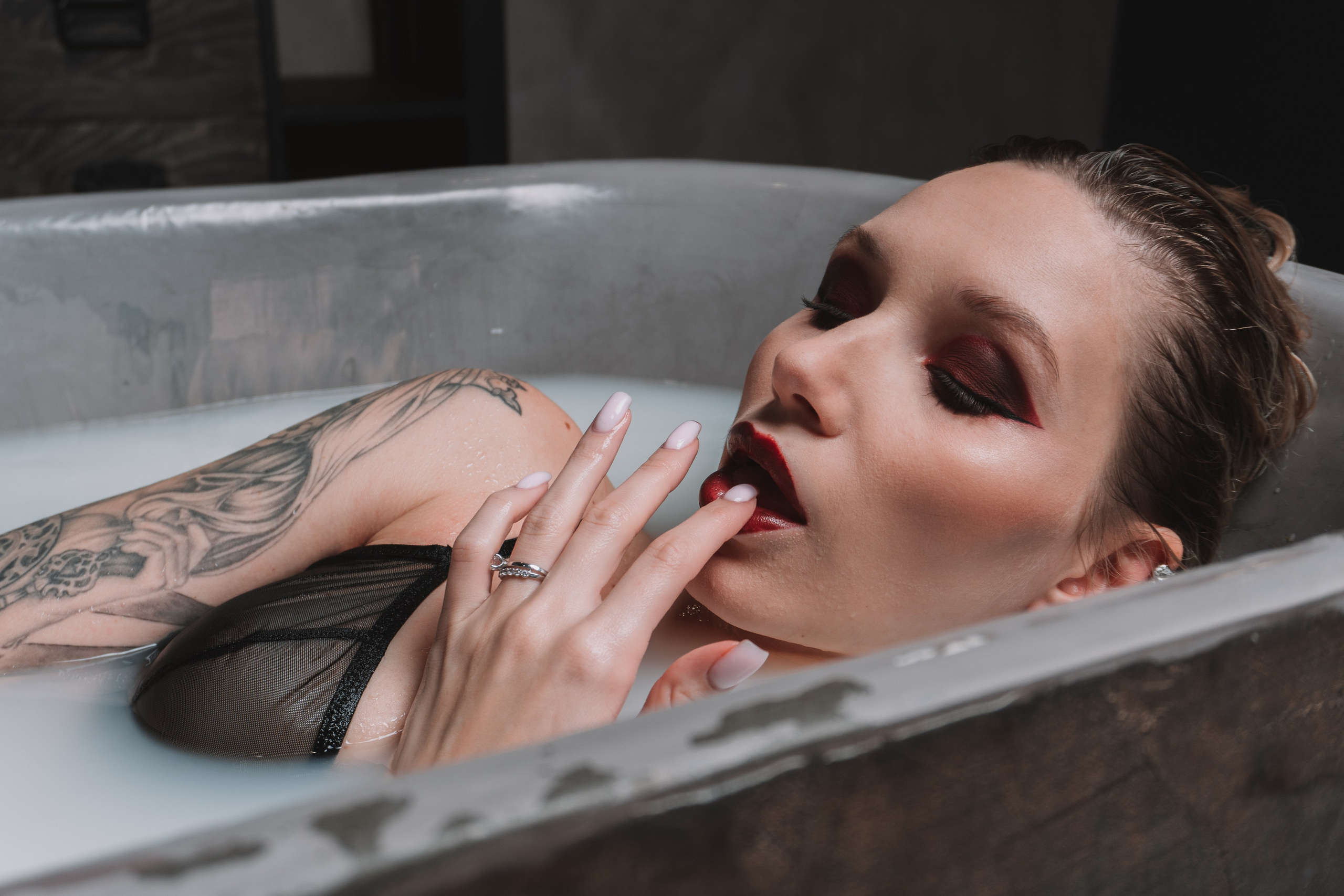 Blonde woman with tattoos lying in a white bath, touching her lips.