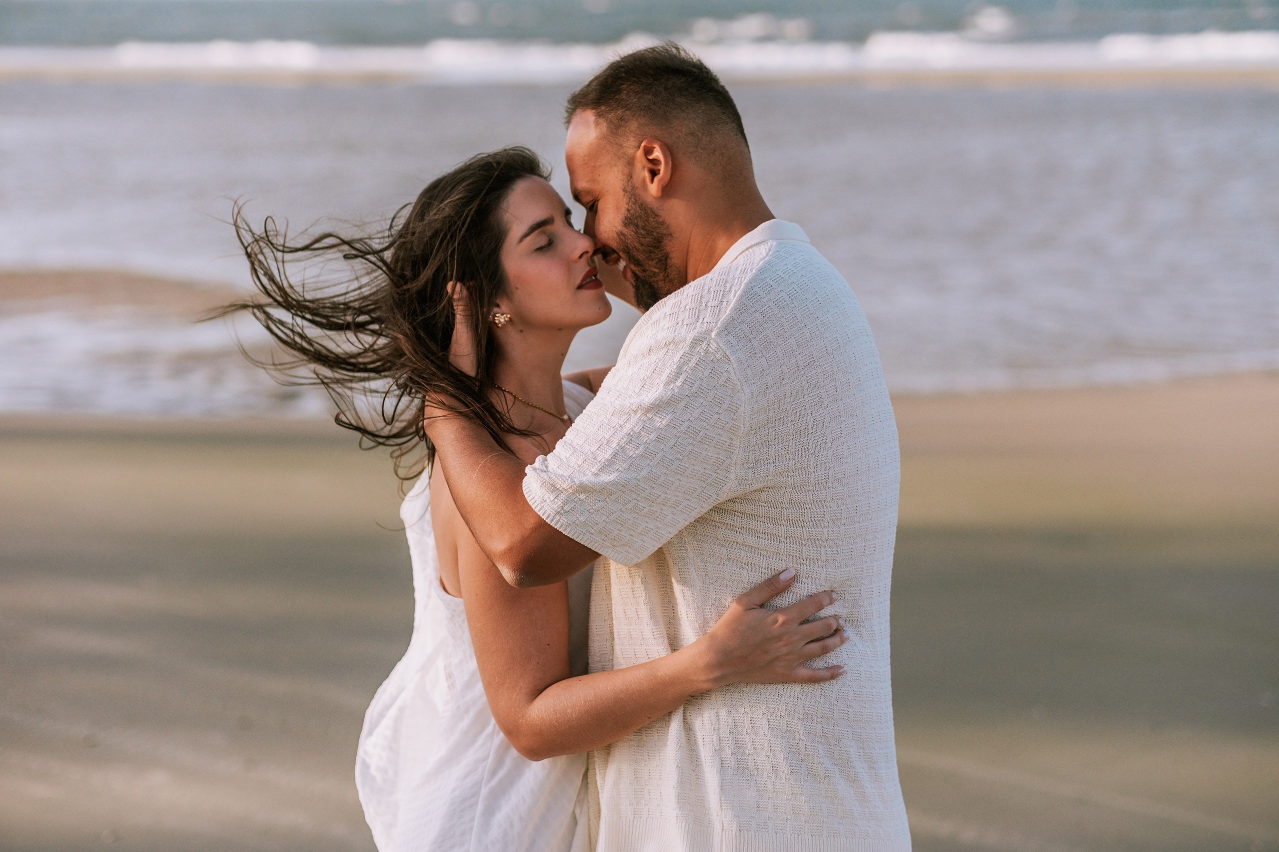 homem e mulher abraçados na praia, os cabelos dela voam ao vento