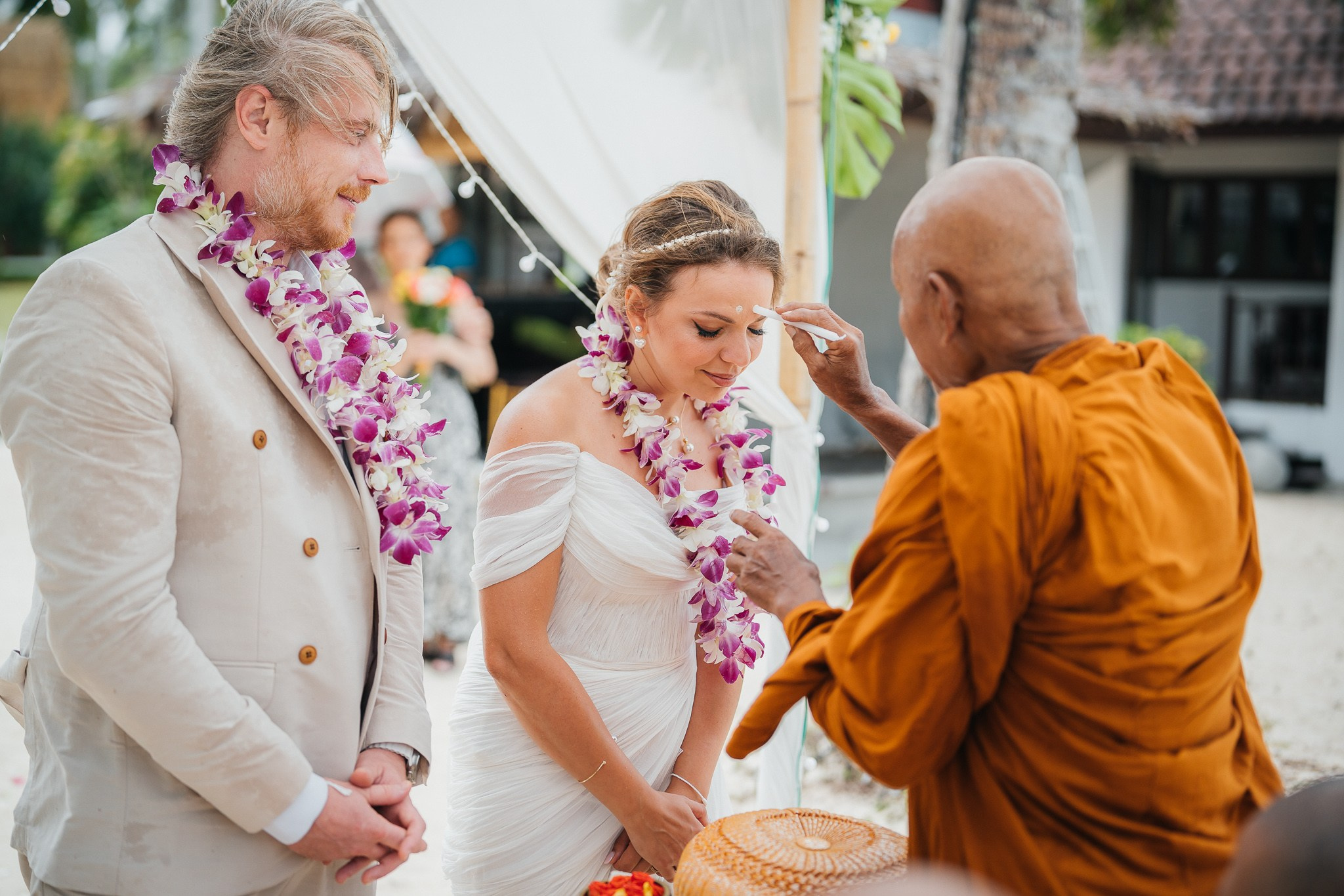 Couple taking part in a traditional Buddhist wedding ceremony on a tropical beach in Koh Samui, Thailand, with monks, flowers, and ocean views.