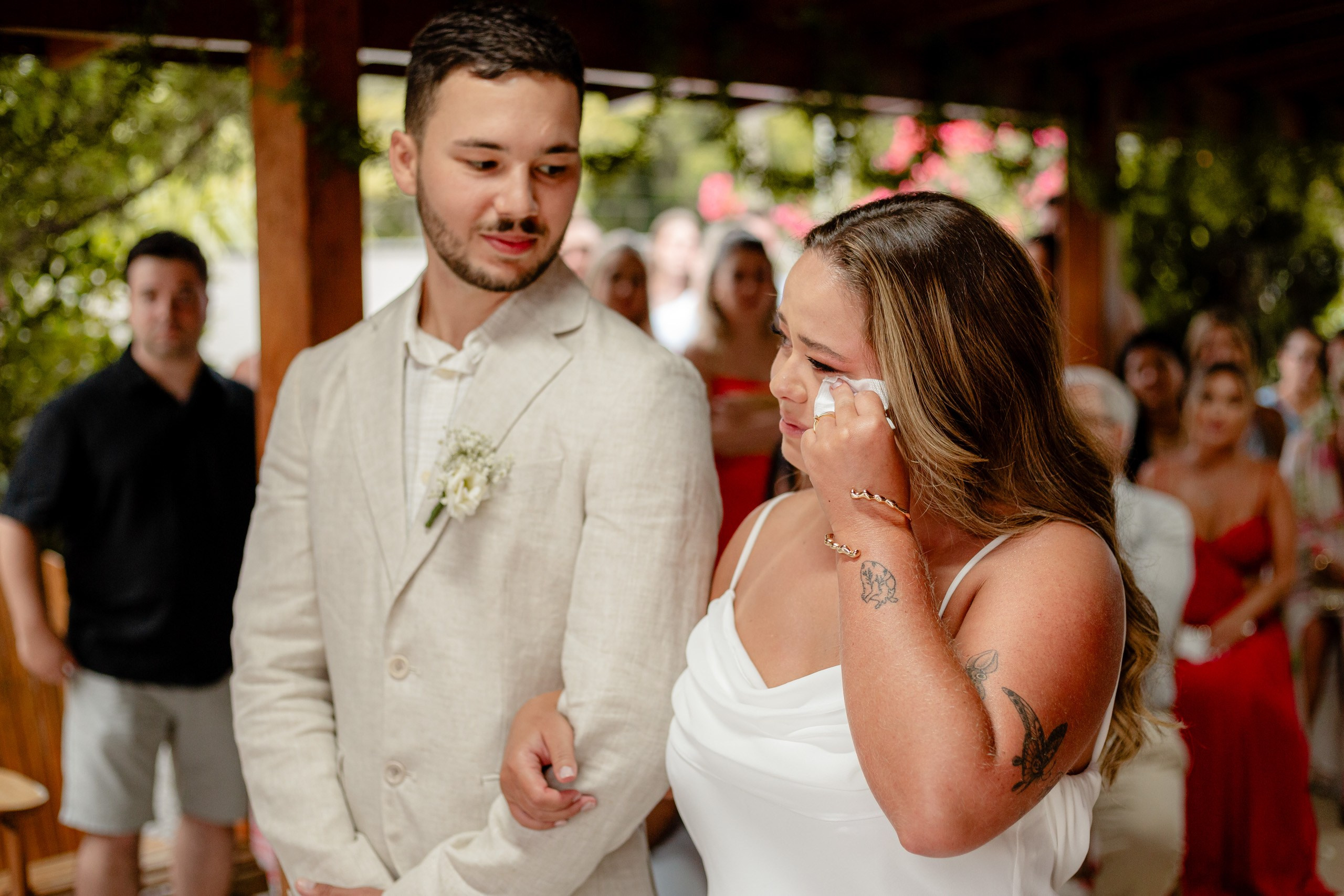 Fotógrafo de casamento em Bombinhas; o noivo observa com carinho a noiva emocionada que limpa uma lágrima durante a cerimônia ao ar livre.