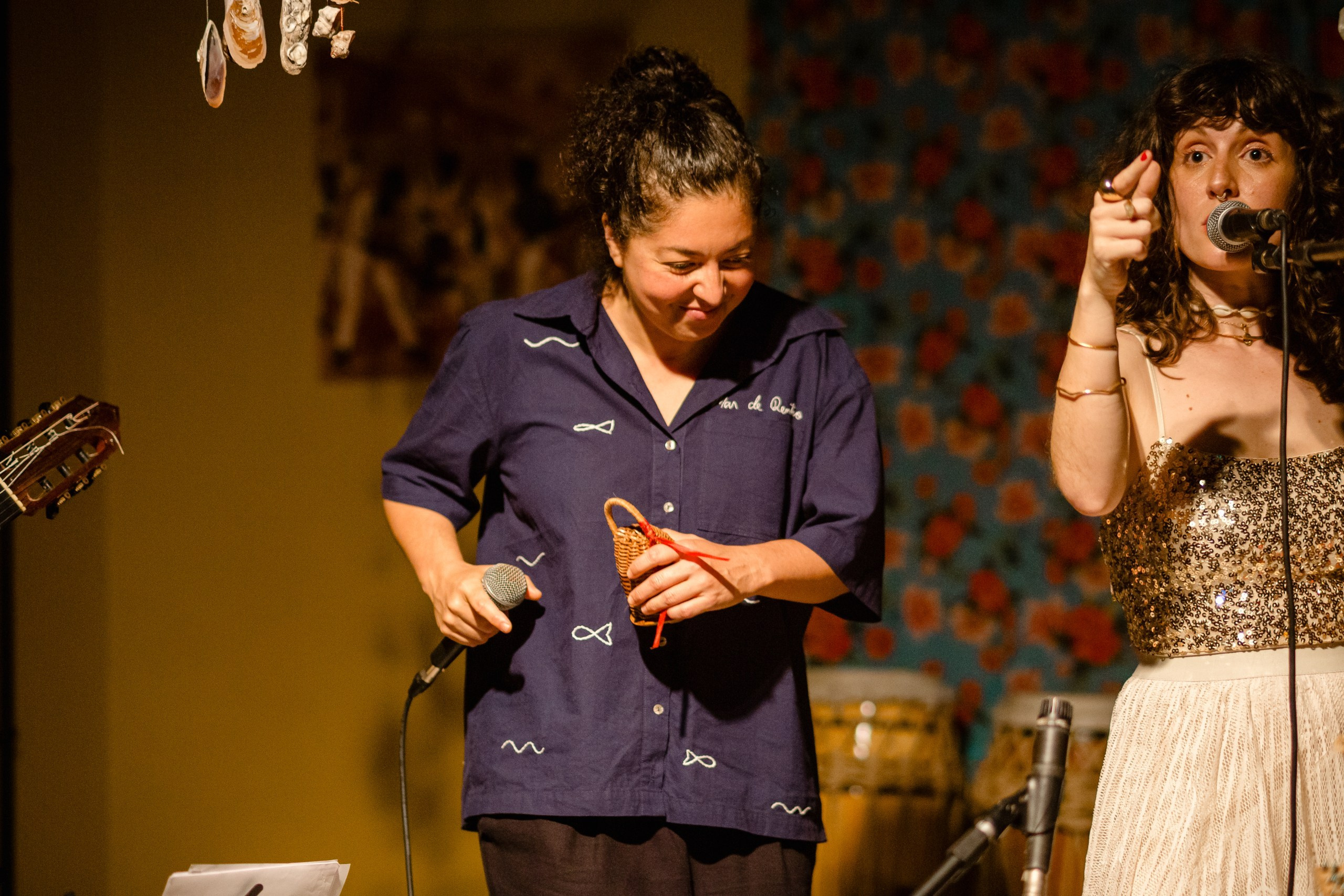 Fotógrafo de show em Balneário Camboriú; a cantora Joana Gaia apontando para o lado enquanto outra artista segura um pequeno instrumento de percussão no palco do bairro da Barra para a divulgação do álbum "Água Brasileira".