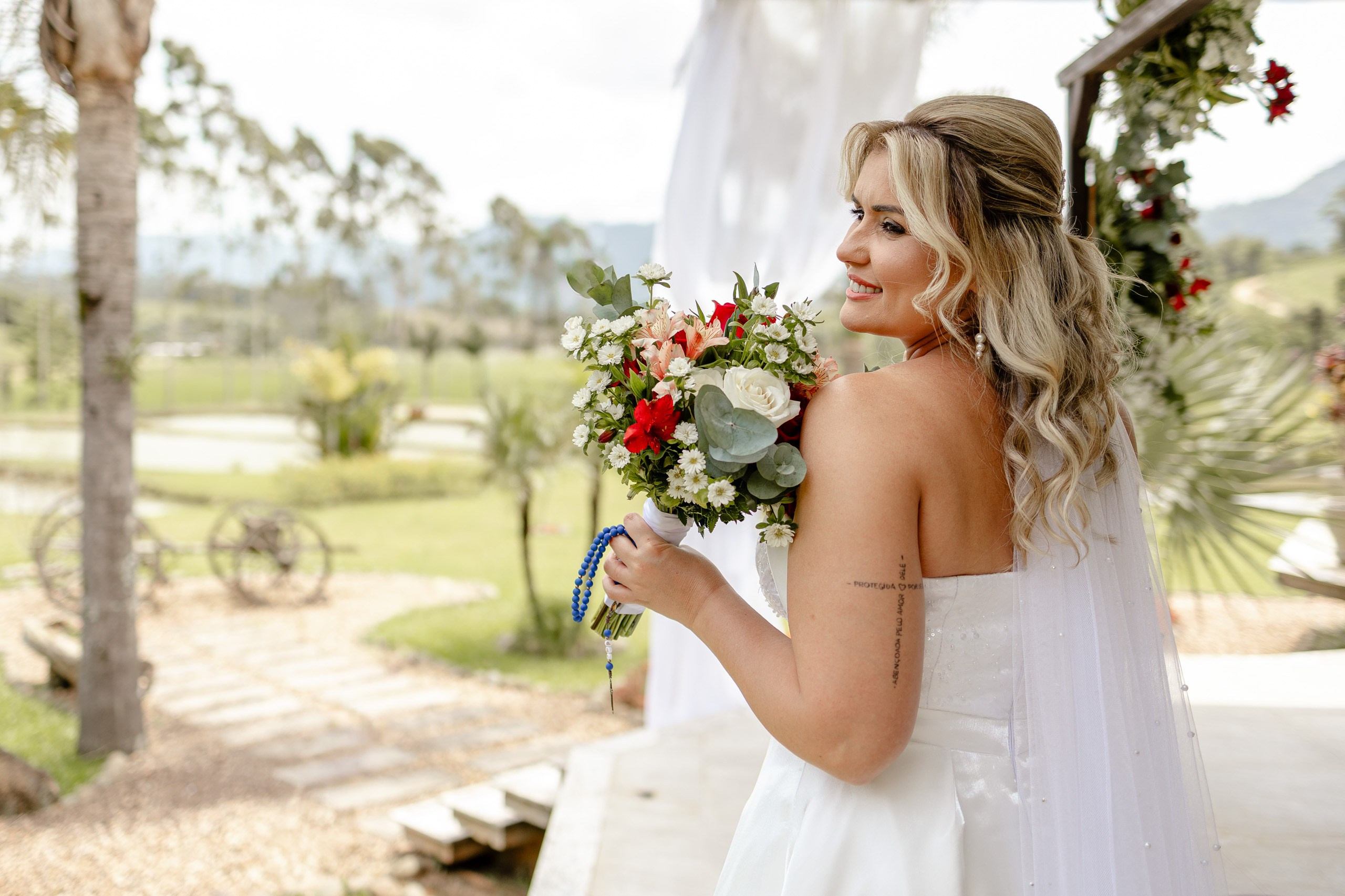 Fotógrafo de casamento em Santa Catarina; noiva sorridente de costas olhando para a paisagem verde com buquê e véu em cerimônia ao ar livre.