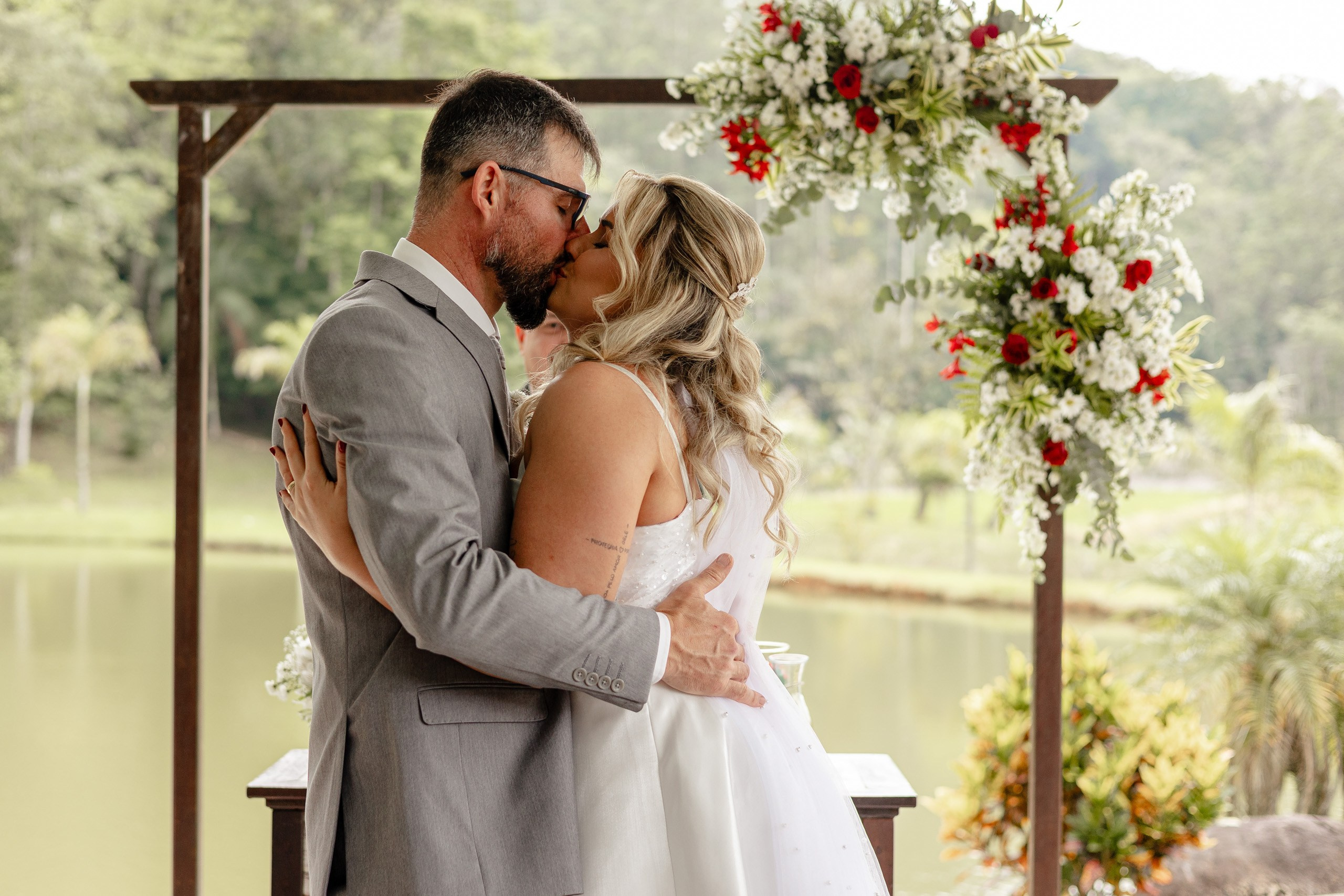 Fotógrafo de casamento em Santa Catarina; beijo dos noivos no altar decorado com flores diante de paisagem natural à beira do lago durante cerimônia no campo.
