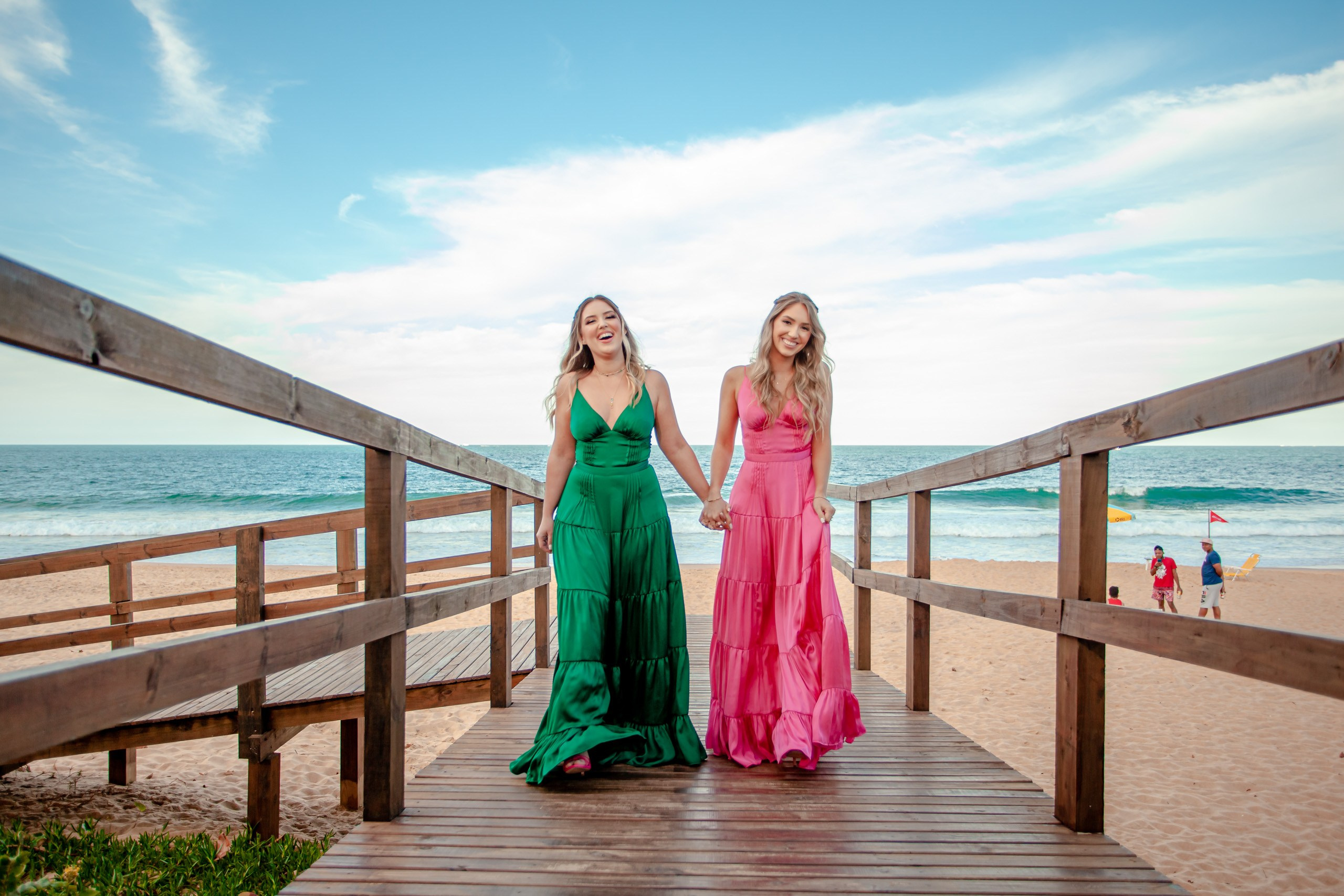 Fotógrafo festa de formatura em Balneário Camboriú; retrato alegre das duas formandas de mãos dadas a caminhar na passarela de madeira com vista para o mar no Bali Bistro Beach.
