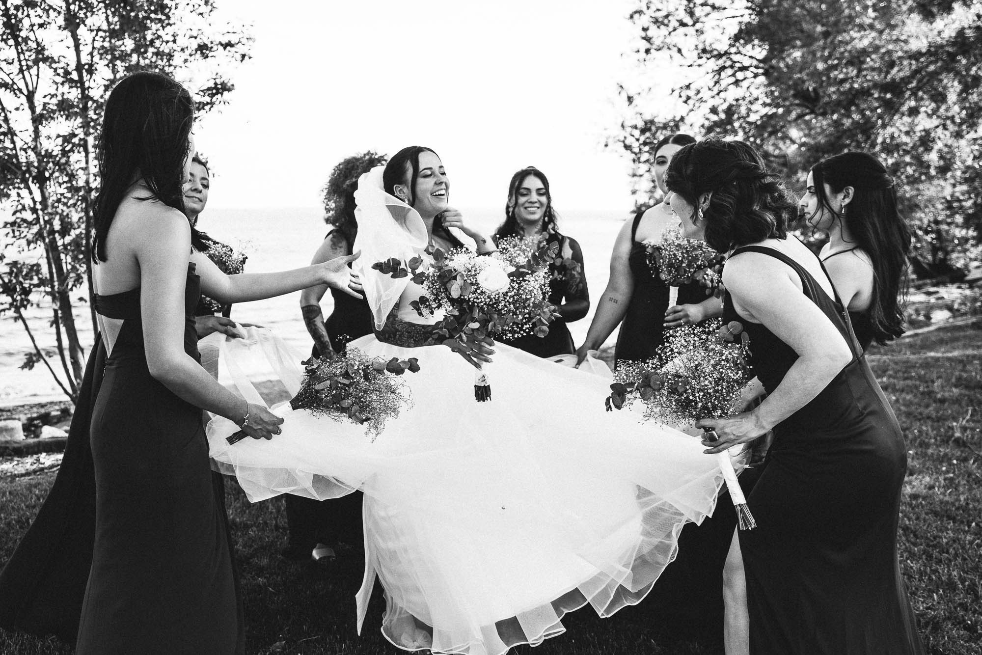 Bridesmaids lift the bride’s dress as they laugh outdoors in black and white.