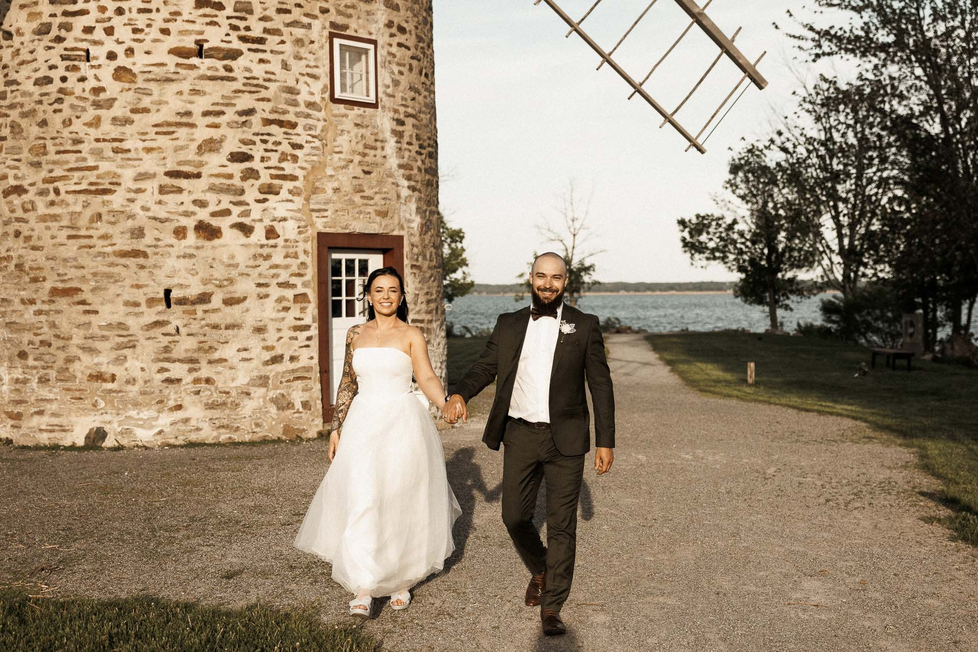 Kendyl and Mikey walk hand in hand beside a stone windmill near the water.