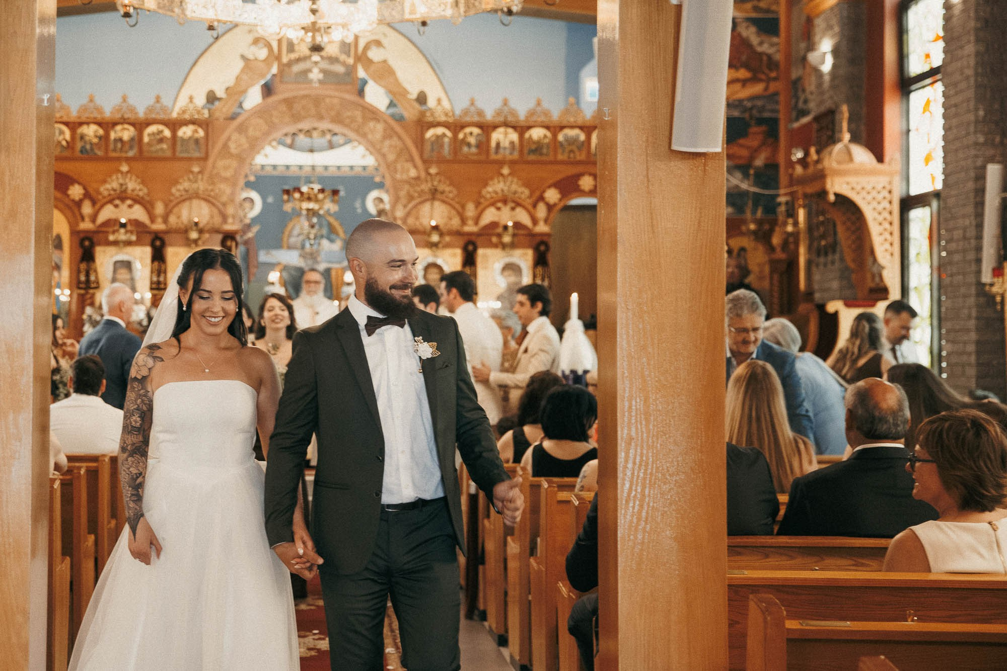The newlyweds walk down the church aisle hand in hand, smiling.