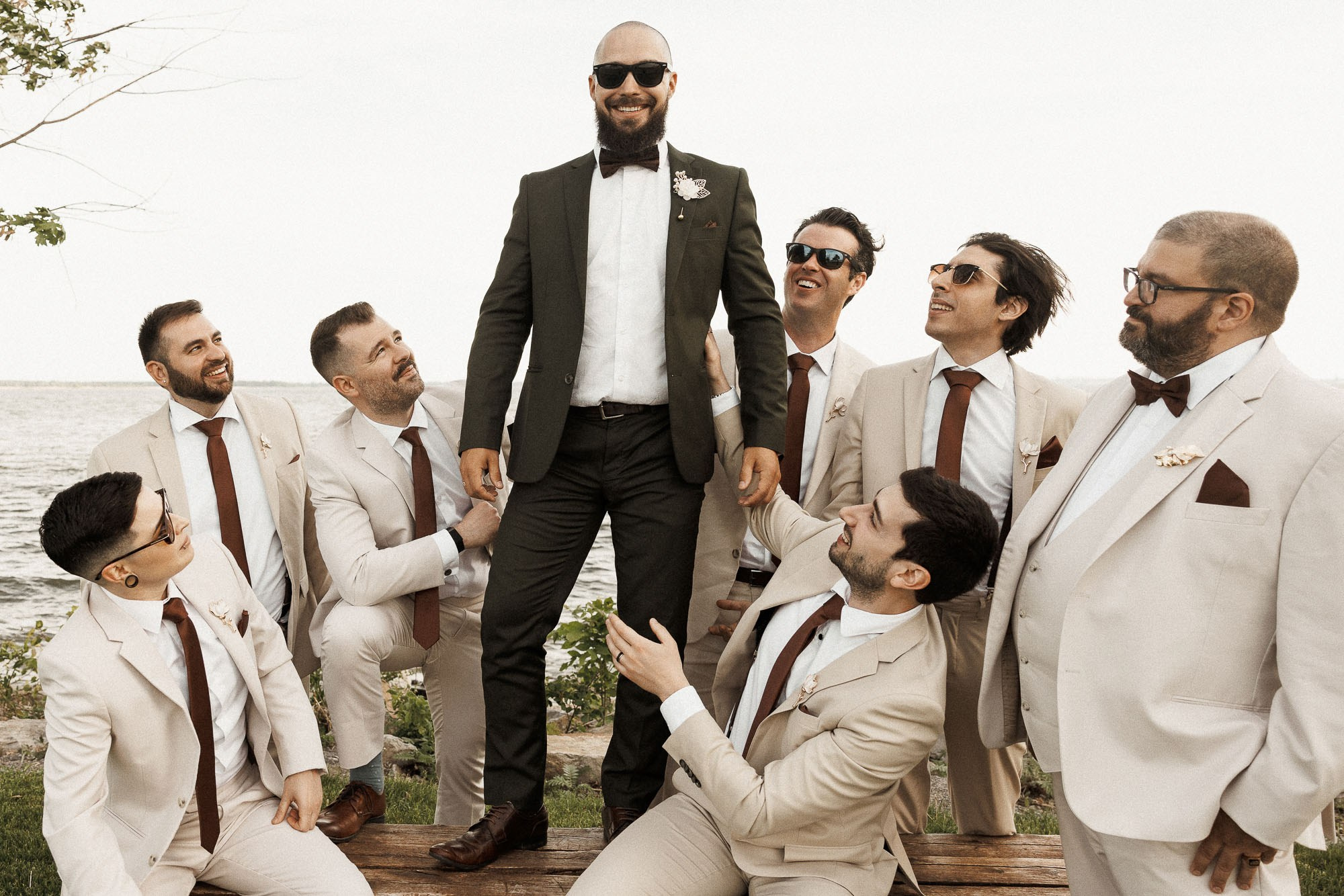 The groom poses with his groomsmen in cream suits by the water.