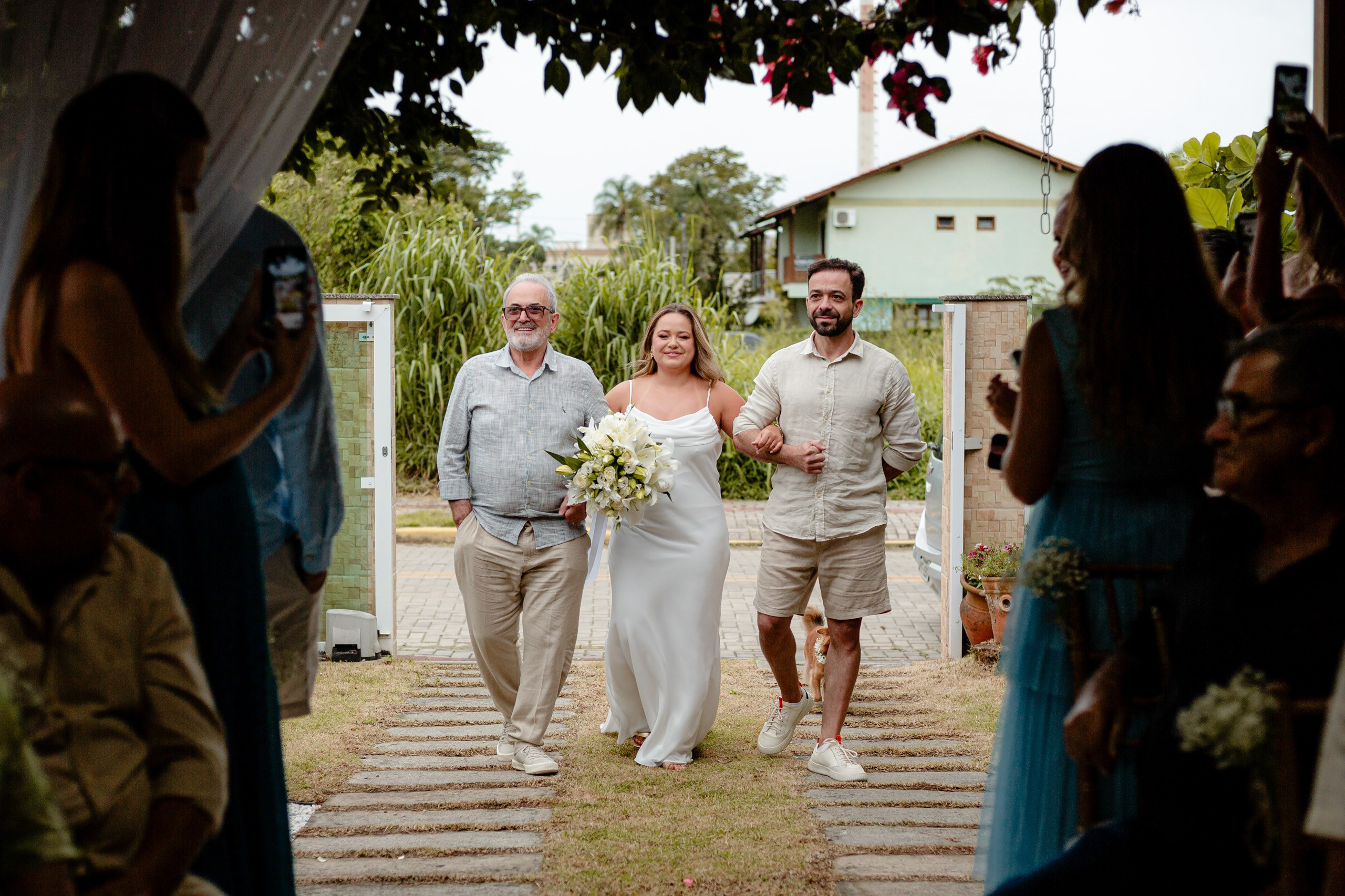 Fotógrafo de casamento em Balneário Camboriú; entrada emocionante da noiva de vestido branco com um buquê de lírios, caminhando por um caminho de jardim entre convidados que registram o momento, acompanhada por dois familiares ao ar livre.