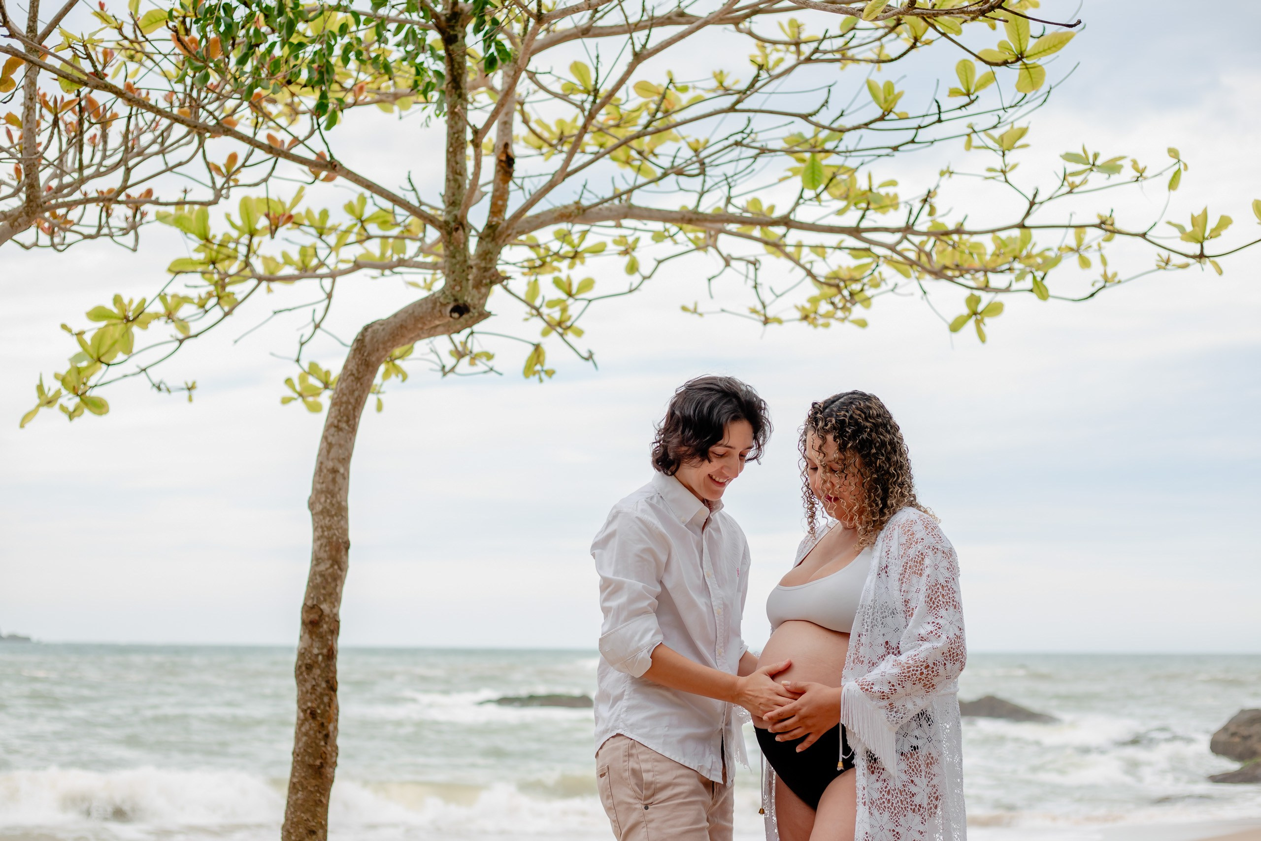 Fotógrafo ensaio gestante em Penha; casal com as mãos apoiadas na barriga em retrato delicado sob árvores na praia, com o mar ao fundo.