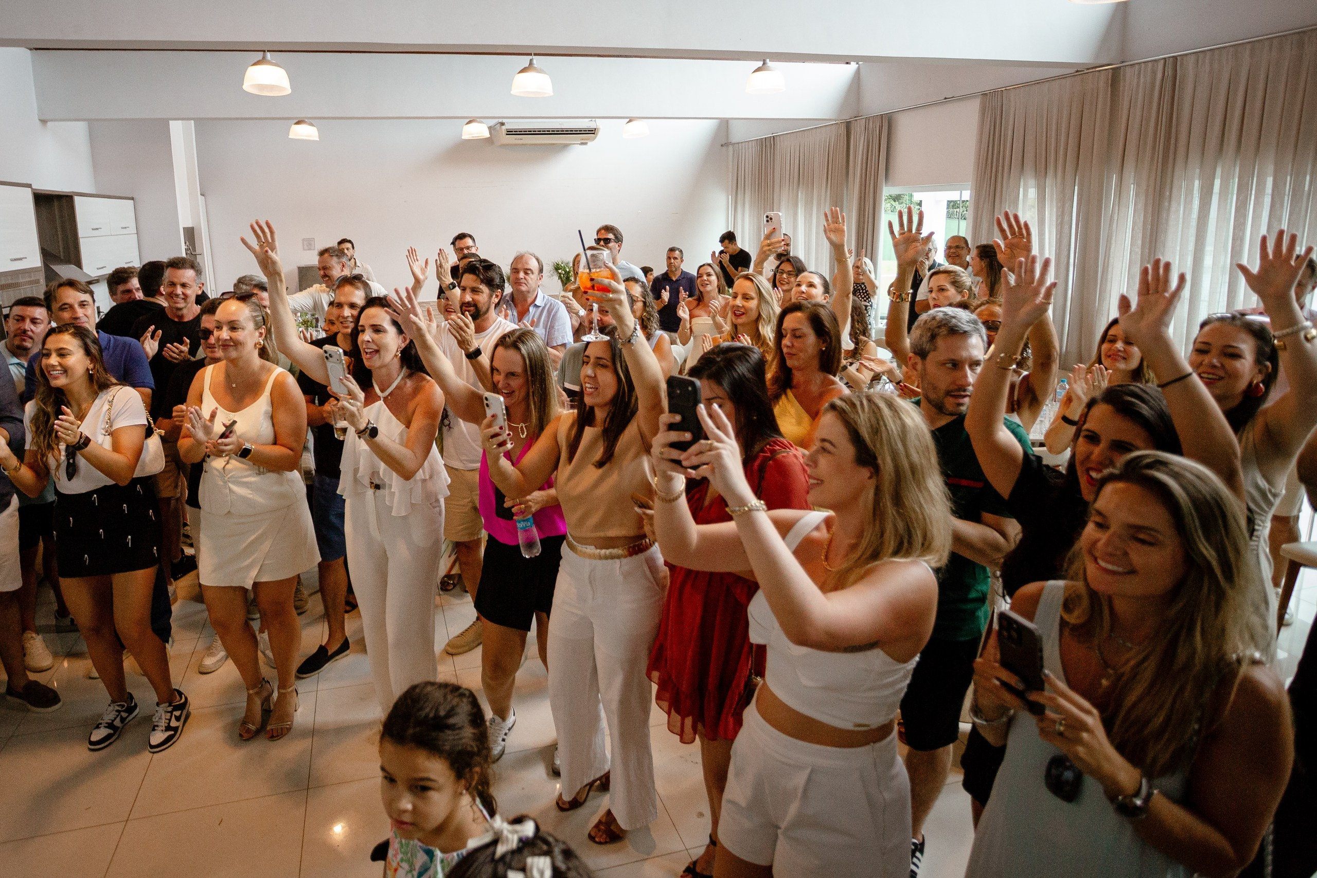 Fotografia de cobertura de festa de aniversário em Itajaí capturando a perspectiva ampla dos convidados a celebrar com entusiasmo, levantar os braços e registrar o momento festivo.
