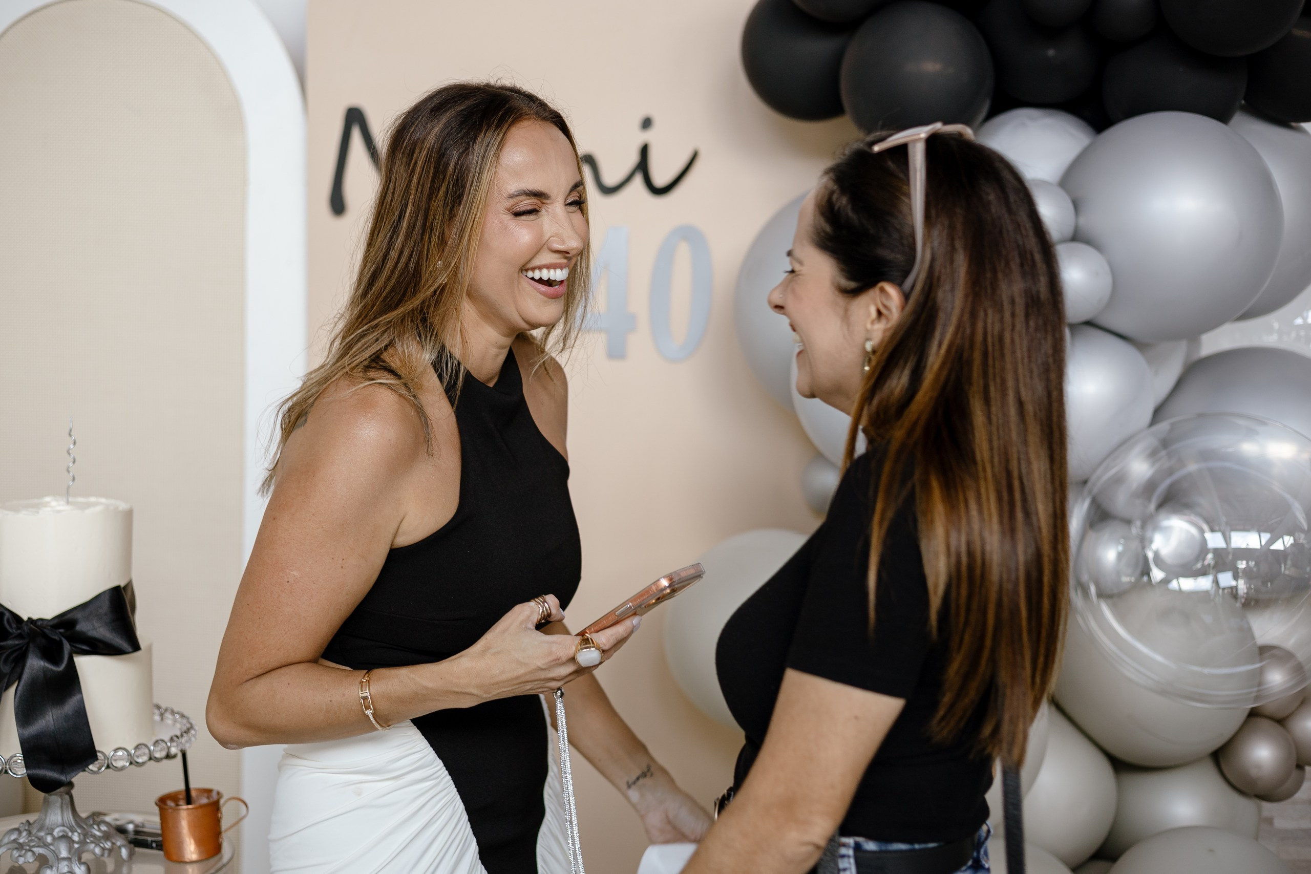 Fotografia de cobertura de festa de aniversário em Itajaí capturando o momento descontraído e de muita gargalhada entre convidada e aniversariante diante do painel decorado com balões.