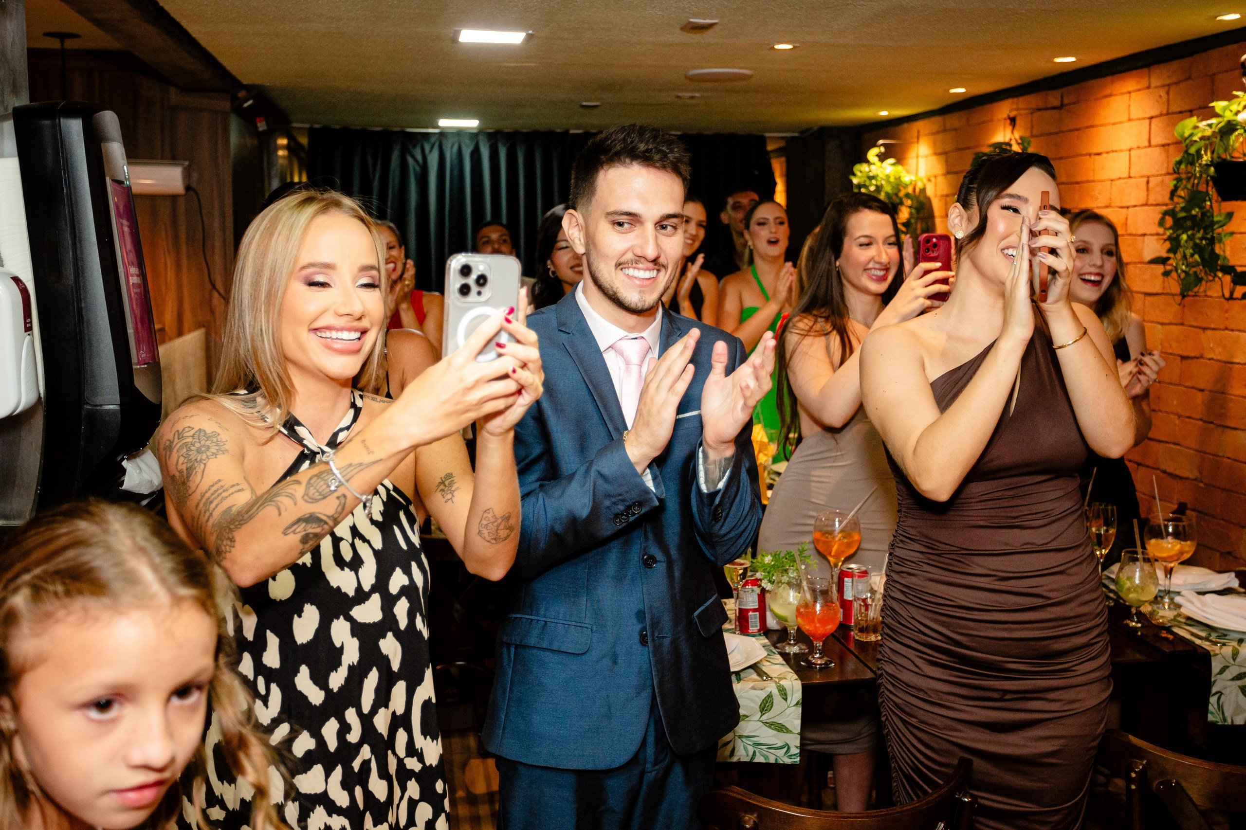 Fotógrafo de formatura em Balneário Camboriú; convidados sorridentes aplaudindo e registrando com o celular durante a festa de comemoração no salão.
