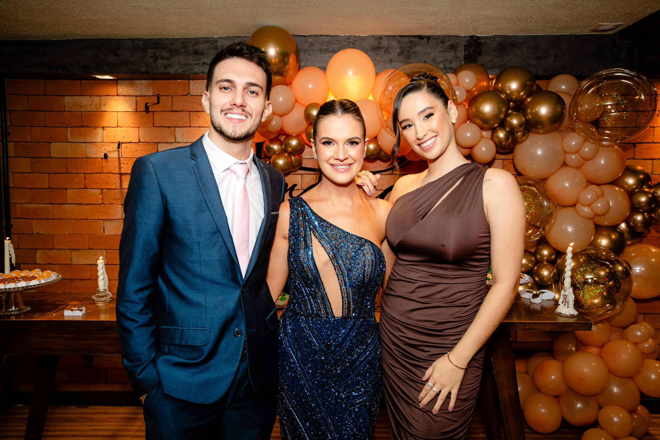 Fotógrafo de formatura em Balneário Camboriú; a formanda posa sorridente ao lado de convidados diante de um painel decorado com balões dourados e laranjas.
