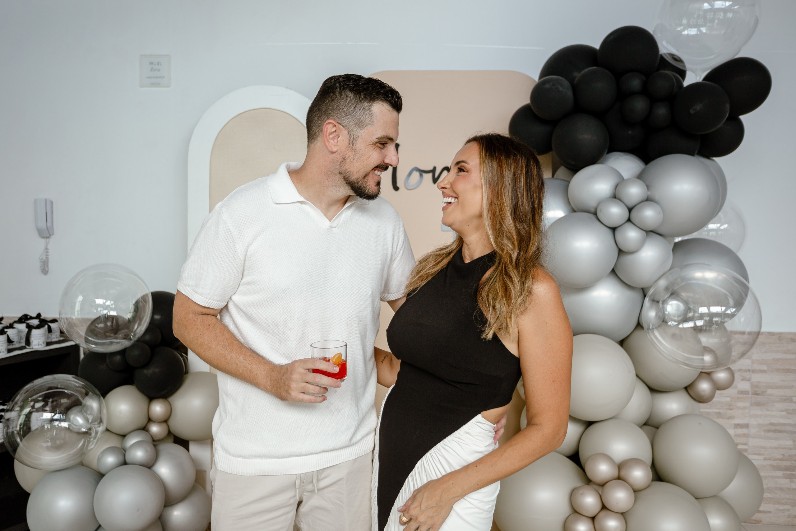 Fotografia de cobertura de festa de aniversário em Itajaí retratando a aniversariante e o marido sorrindo um para o outro em um momento romântico e descontraído diante do painel com balões.