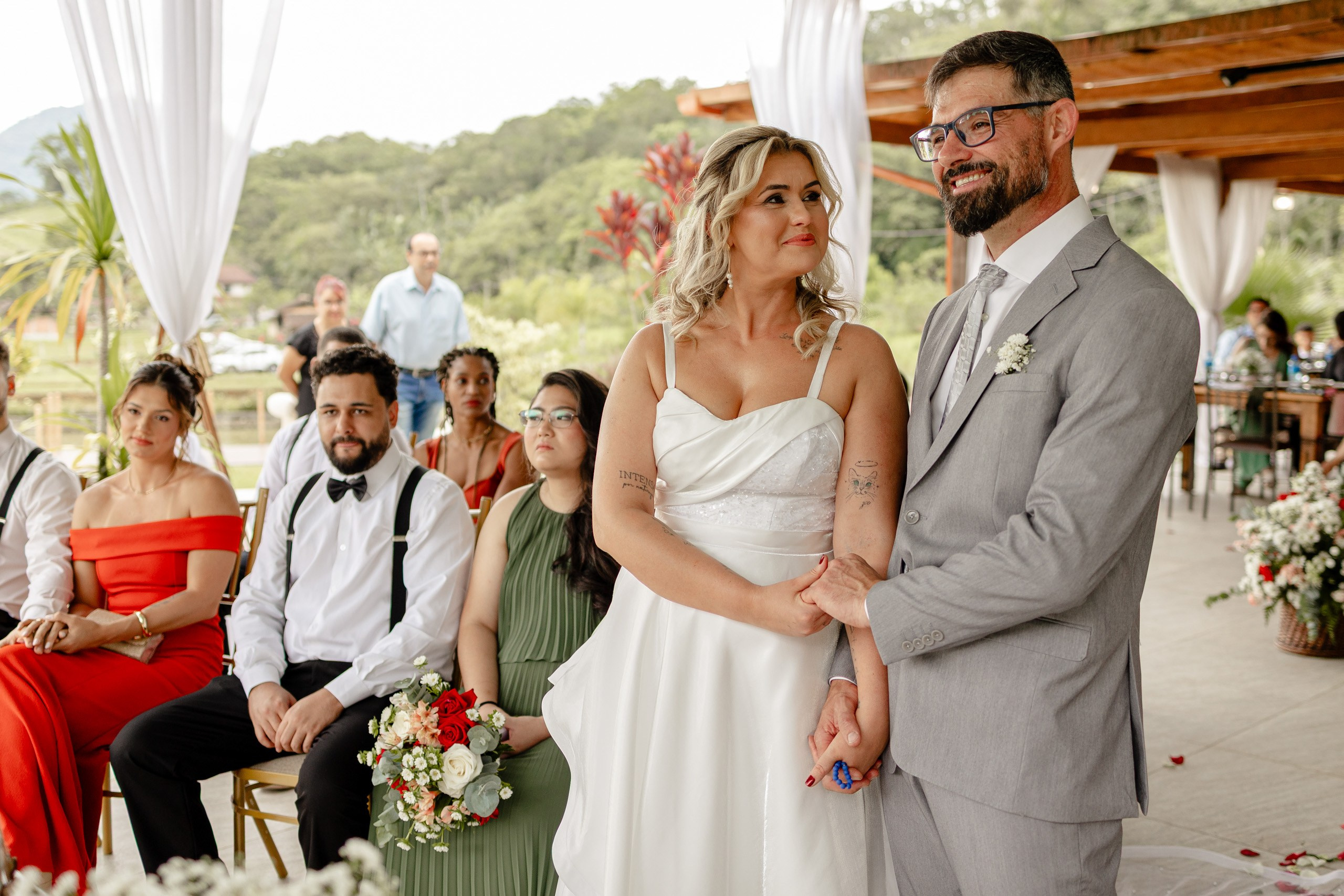Fotógrafo de casamento em Santa Catarina; noivos de mãos dadas emocionados no altar, com padrinhos e madrinhas sentados ao fundo durante cerimônia no campo.