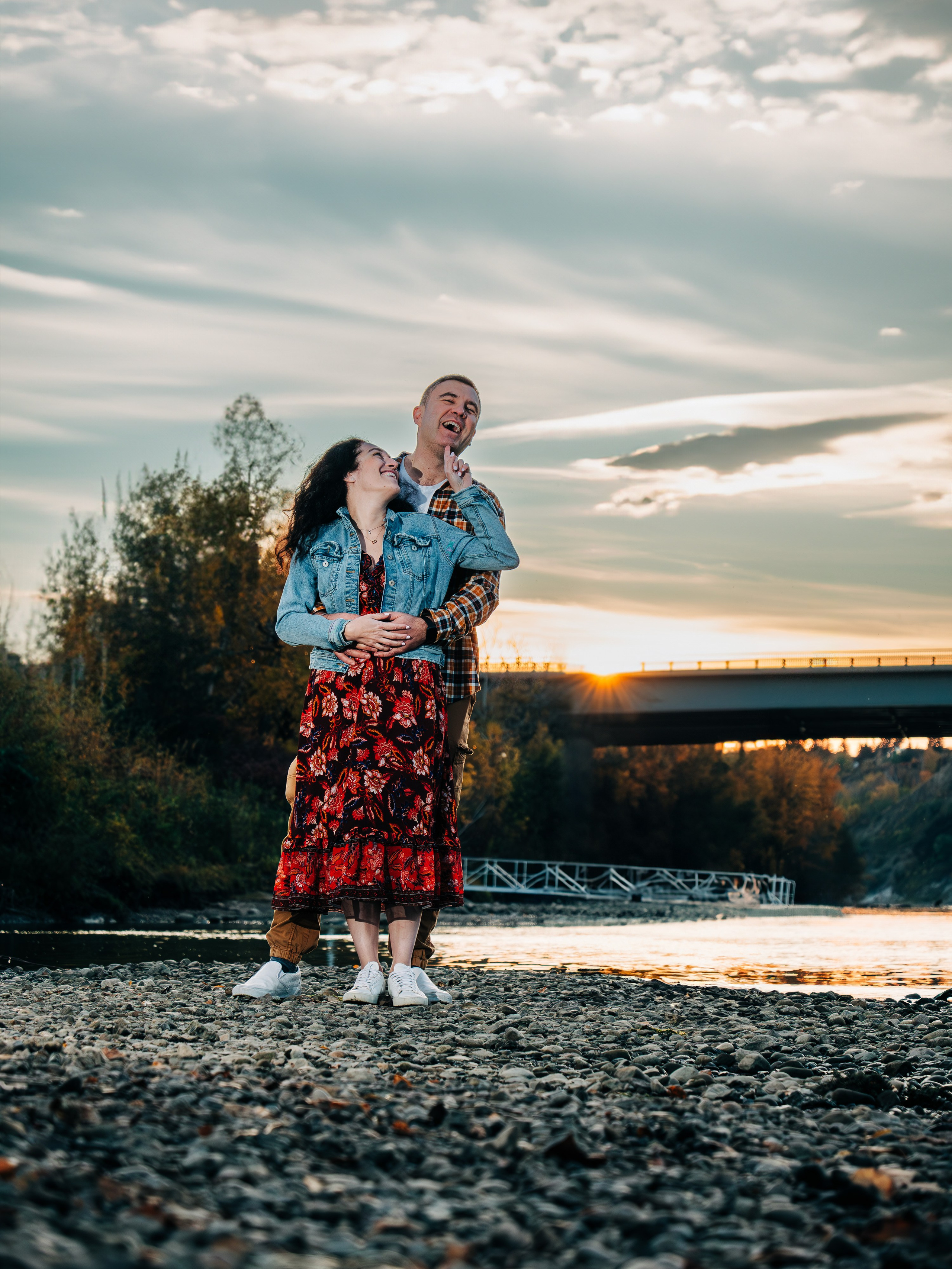 Professional couples photography by Dimitri Studio. Happy couple standing on a rocky riverbank at sunset with a bridge in the background, Spruce Grove. Book at dimitristudio.com