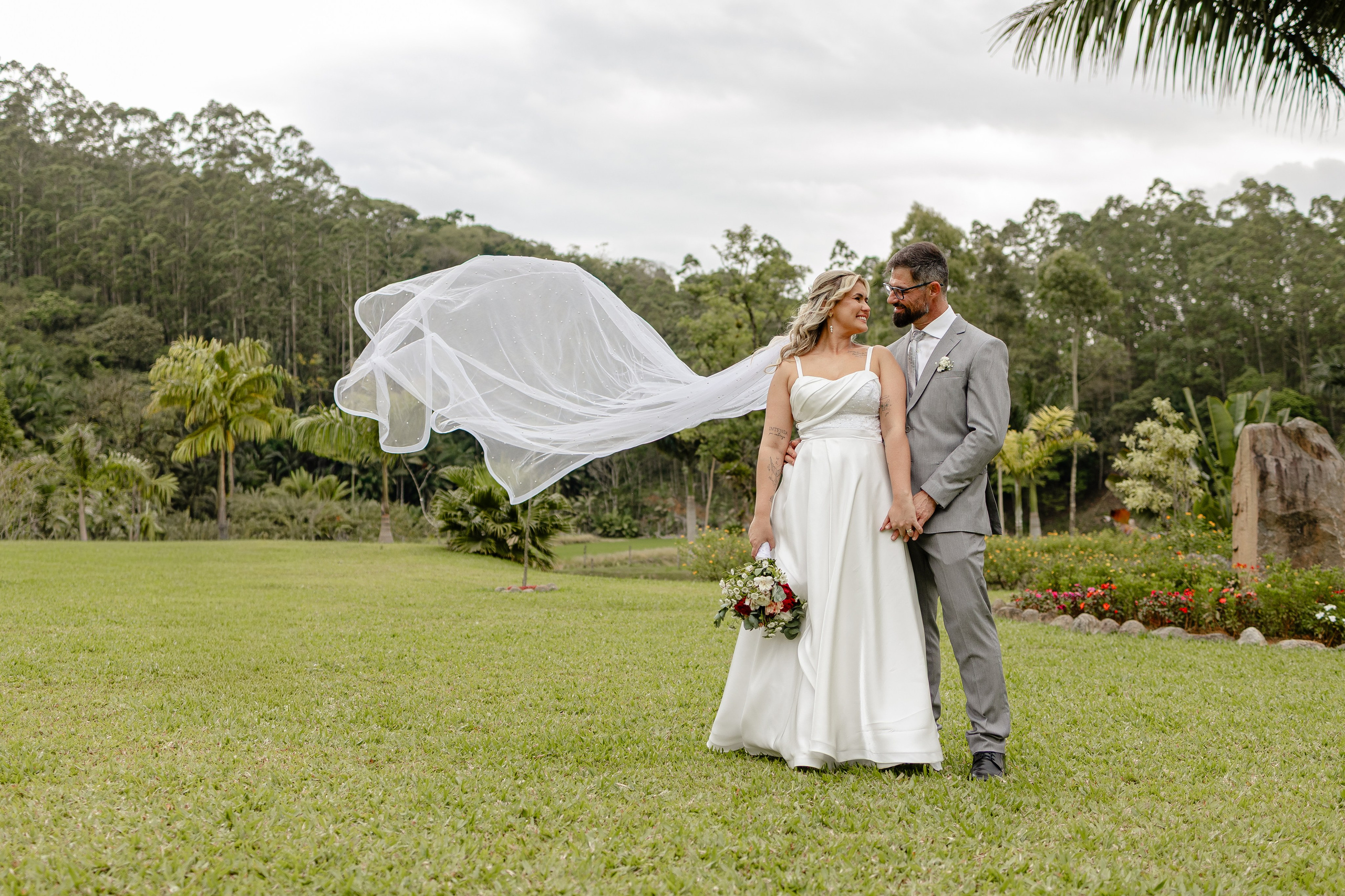 Fotógrafo de casamento em Balneário Camboriú; retrato romântico e amplo de recém-casados em um vasto gramado verde ao ar livre, com o noivo de terno cinza, a noiva de vestido branco a segurar um buquê e o véu flutuando artisticamente ao vento.