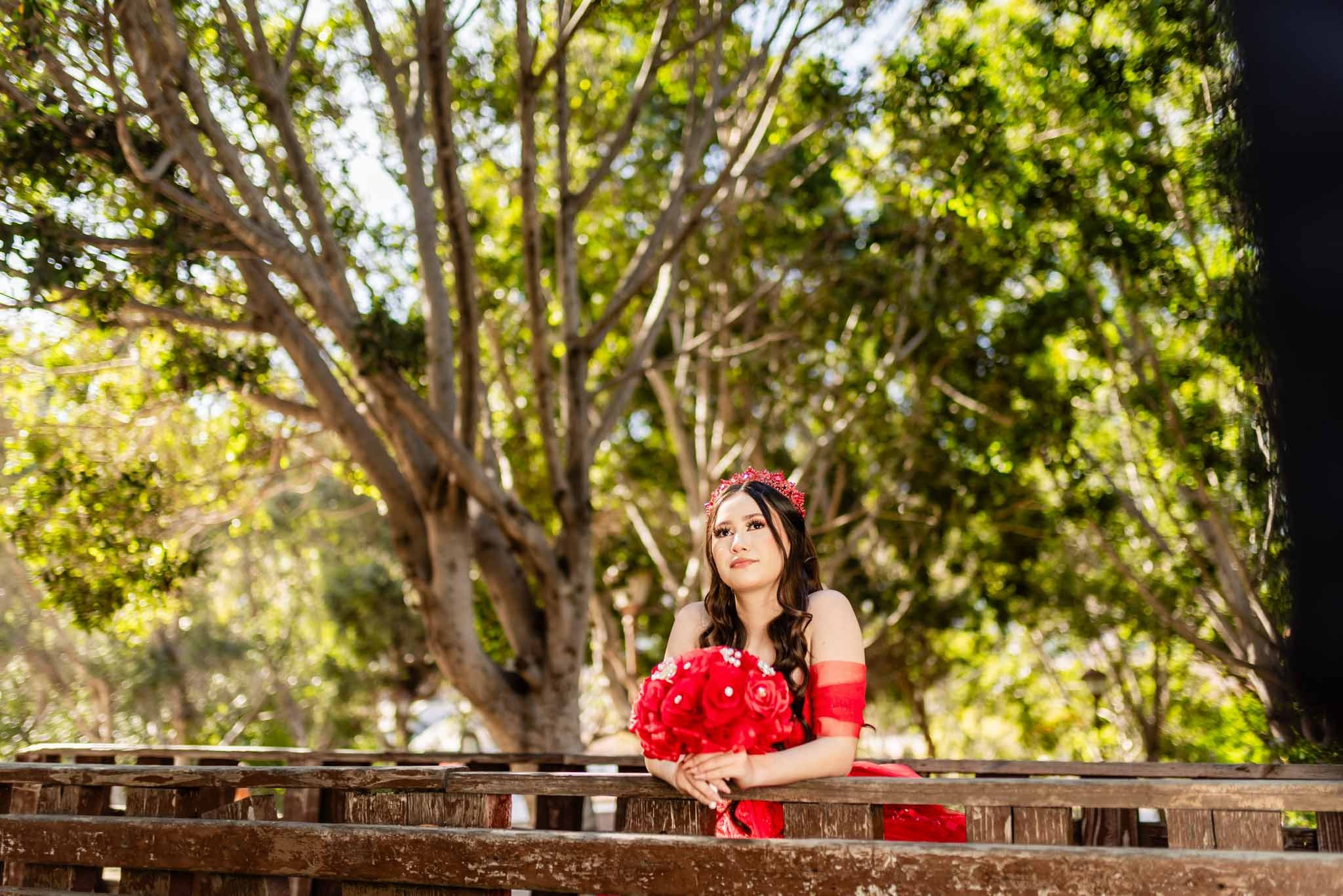 Quinceanera en Parque Hacienda Aguacaliente Tijuana apoyada en barandal con fondo de arboles vestido rojo sesion fotografica exterior