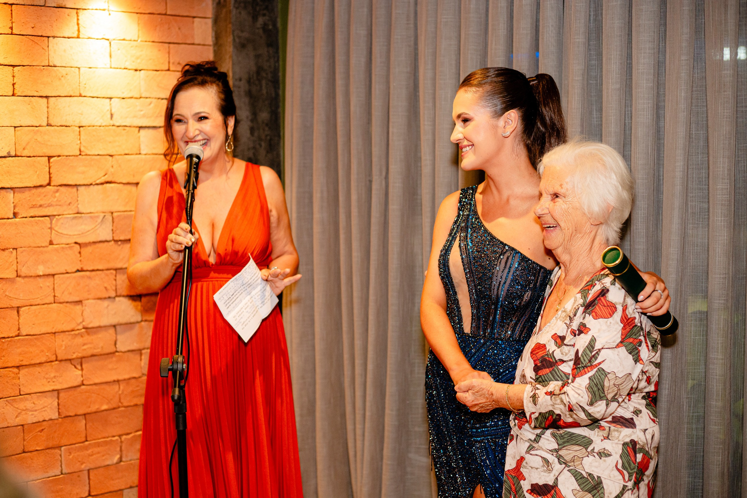 Fotógrafo de formatura em Balneário Camboriú; mulher com vestido vermelho discursa ao microfone enquanto a formanda e uma convidada sorriem juntas durante a festa do evento.