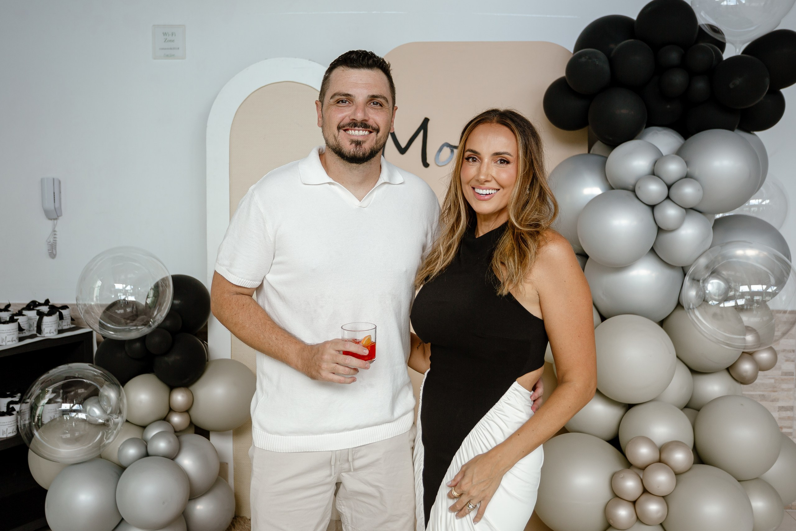 Fotografia de cobertura de festa de aniversário em Itajaí retratando a aniversariante sorridente ao lado do marido, que segura um copo com drink, em frente ao painel decorado com balões.
