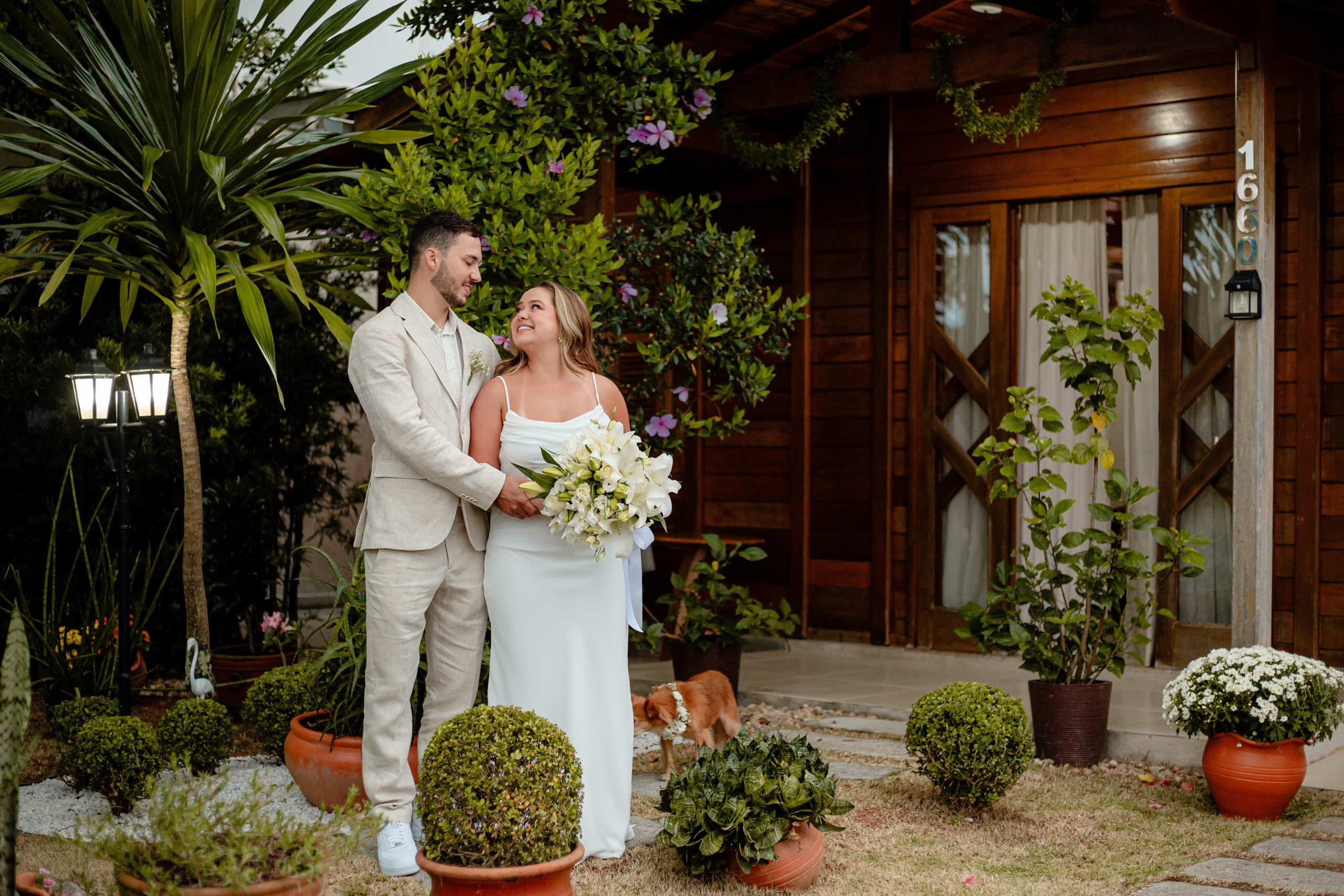 Fotógrafo de casamento em Balneário Camboriú; retrato romântico e aberto do casal a olhar um para o outro em frente a uma casa de madeira com jardim, acompanhados pelo seu cãozinho e com a noiva segurando um buquê de flores brancas.