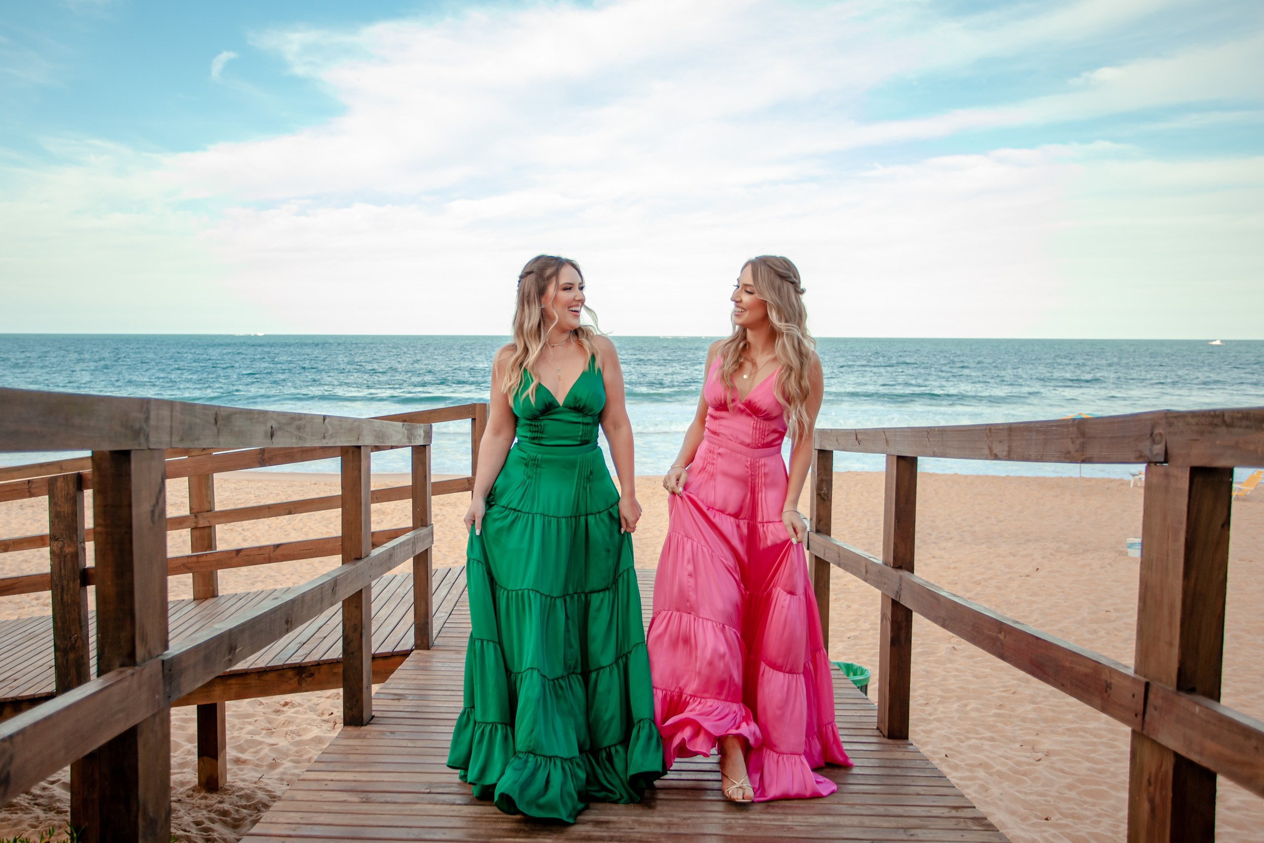Fotógrafo festa de formatura em Balneário Camboriú; retrato externo elegante e sorridente das duas formandas em passarela de madeira na praia do Bali Bistro Beach.