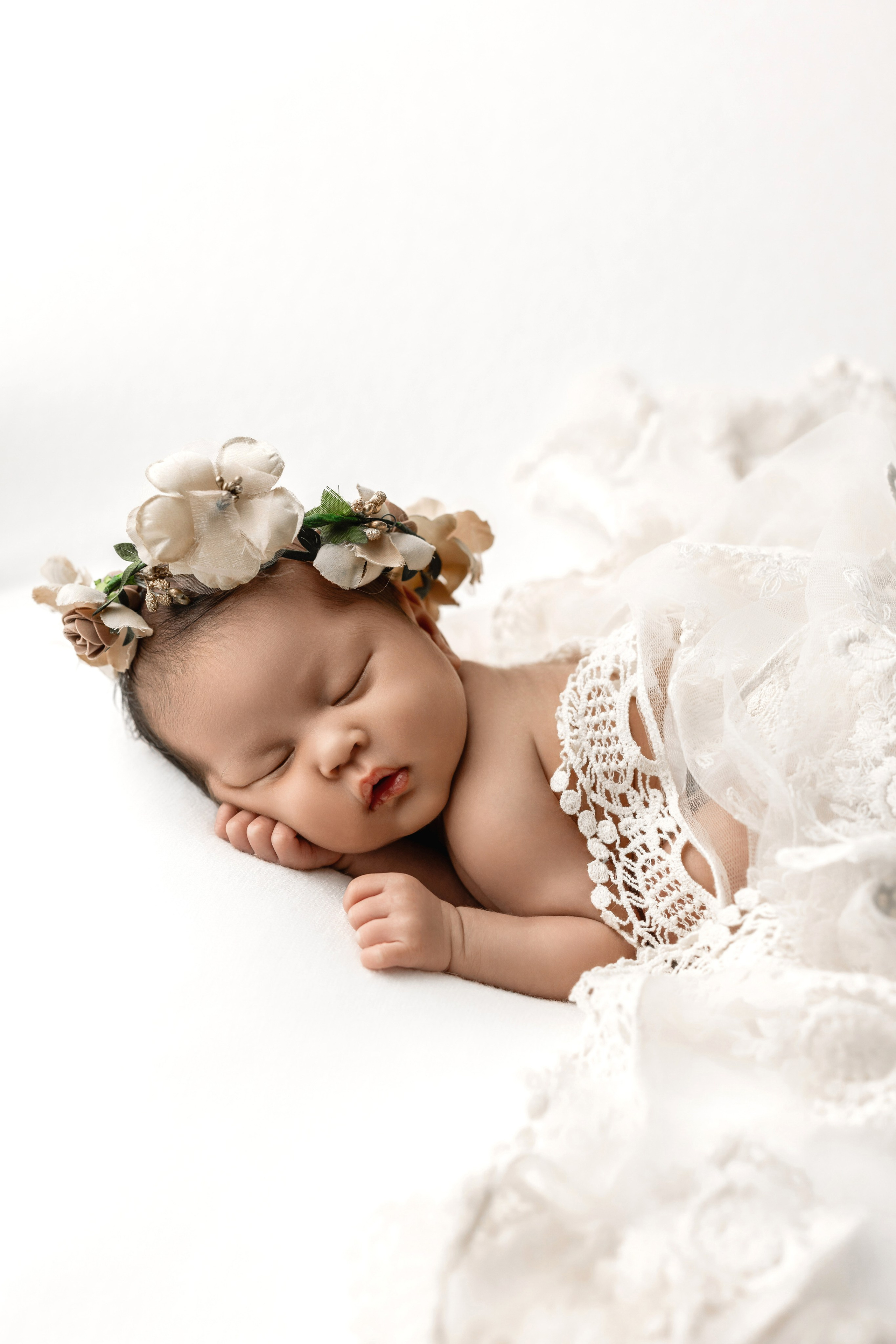 Newborn baby girl sleeping on a white background, wearing a floral crown and wrapped in a delicate lace blanket — fine art newborn photography with soft neutral tones.