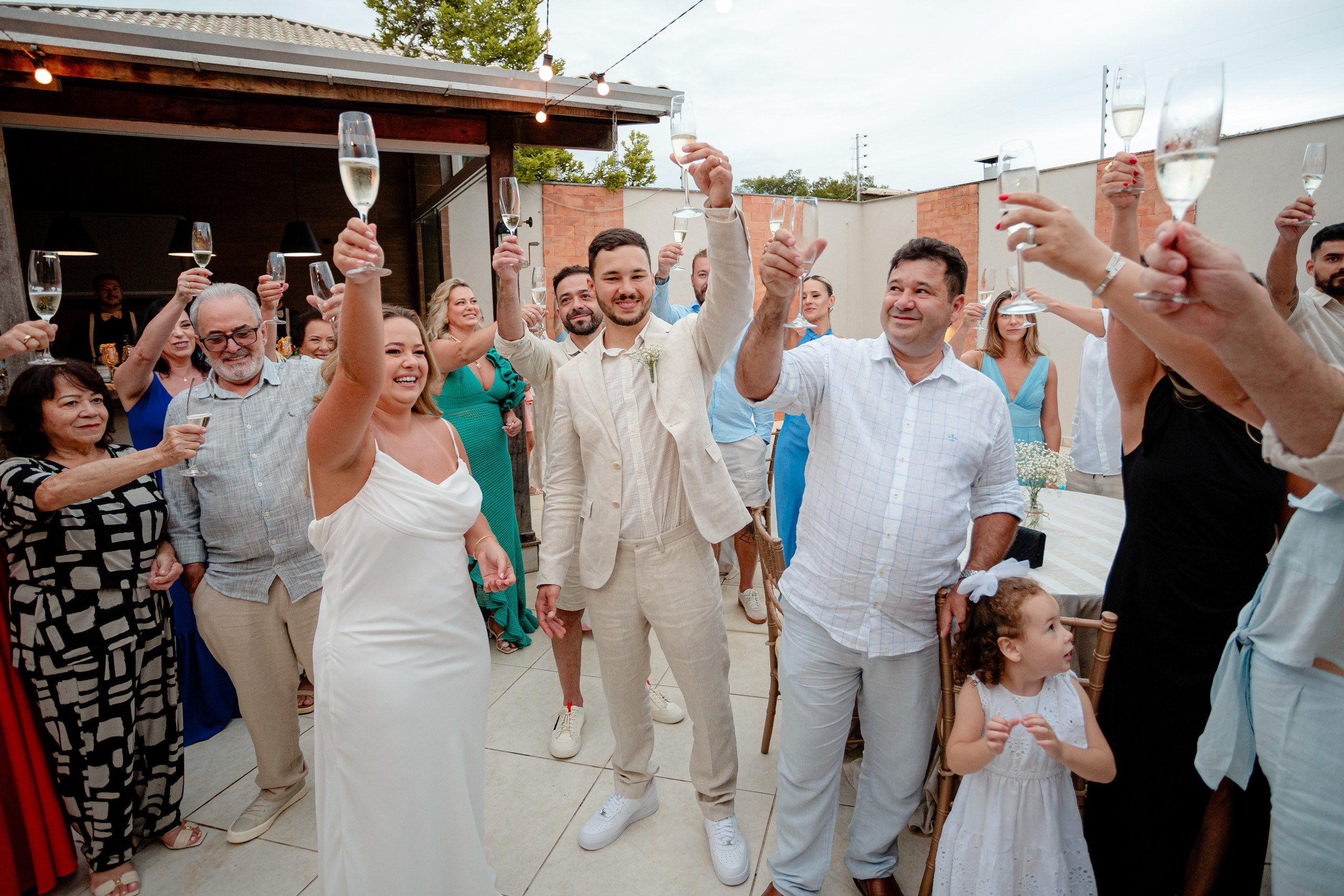 Fotógrafo de casamento em Bombinhas; os noivos erguem taças para um brinde festivo com os convidados durante a comemoração ao ar livre.