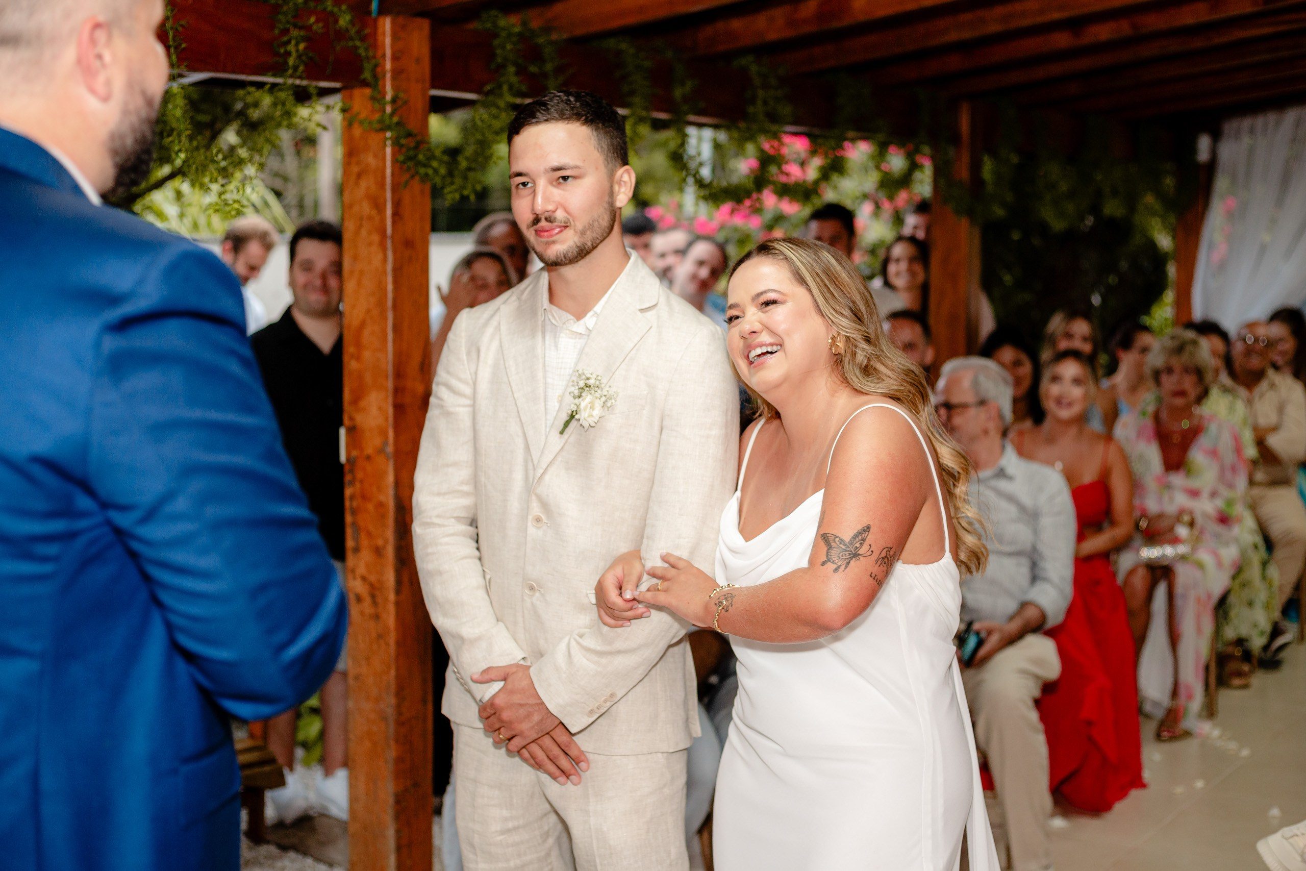 Fotógrafo de casamento em Bombinhas; o noivo e a noiva riem alegremente de braço dado durante a cerimônia sob a estrutura de madeira.