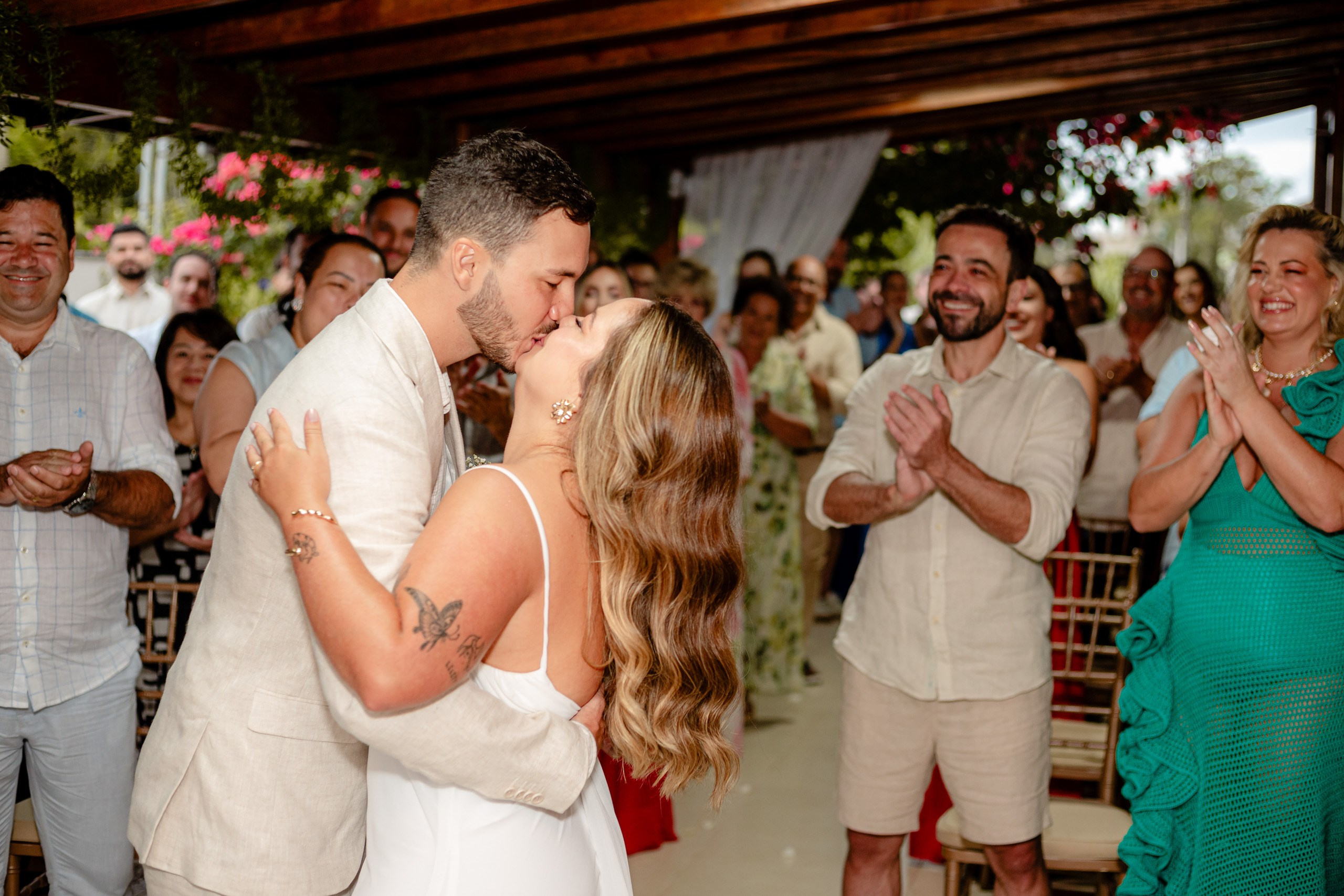 Fotógrafo de casamento em Bombinhas; o noivo e a noiva dão o beijo oficial do matrimônio, celebrados e aplaudidos com entusiasmo pelos convidados presentes.