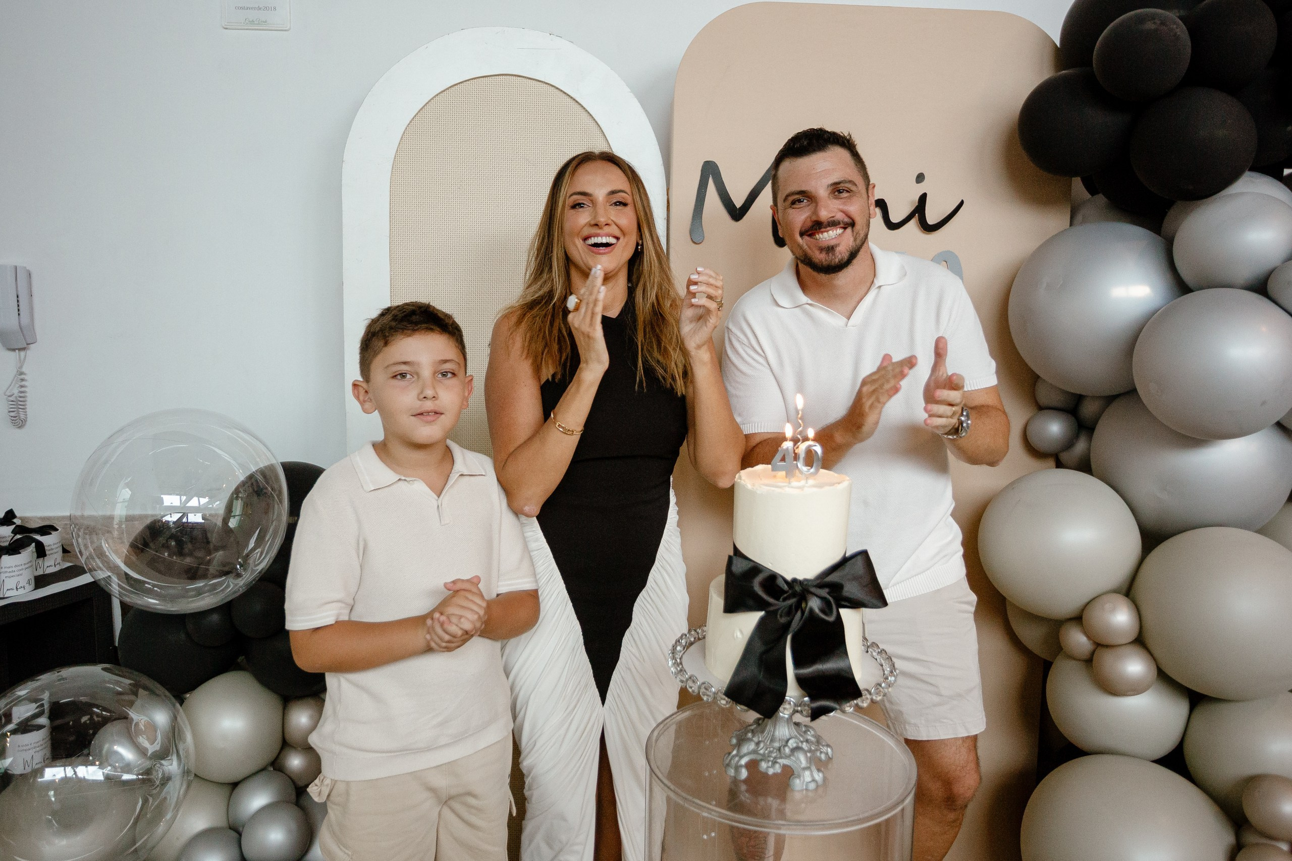 Fotografia de cobertura de festa de aniversário em Itajaí retratando a família reunida em torno do bolo de 40 anos, com a aniversariante sorrindo e cantando os parabéns ao lado do marido e do filho.