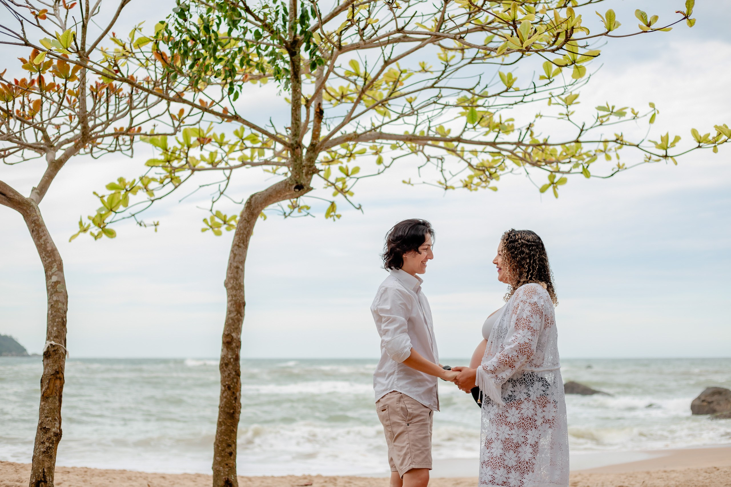 Fotógrafo ensaio gestante em Penha; casal de mãos dadas sob a sombra de árvores na praia, com o mar ao fundo em um retrato afetivo e documental.