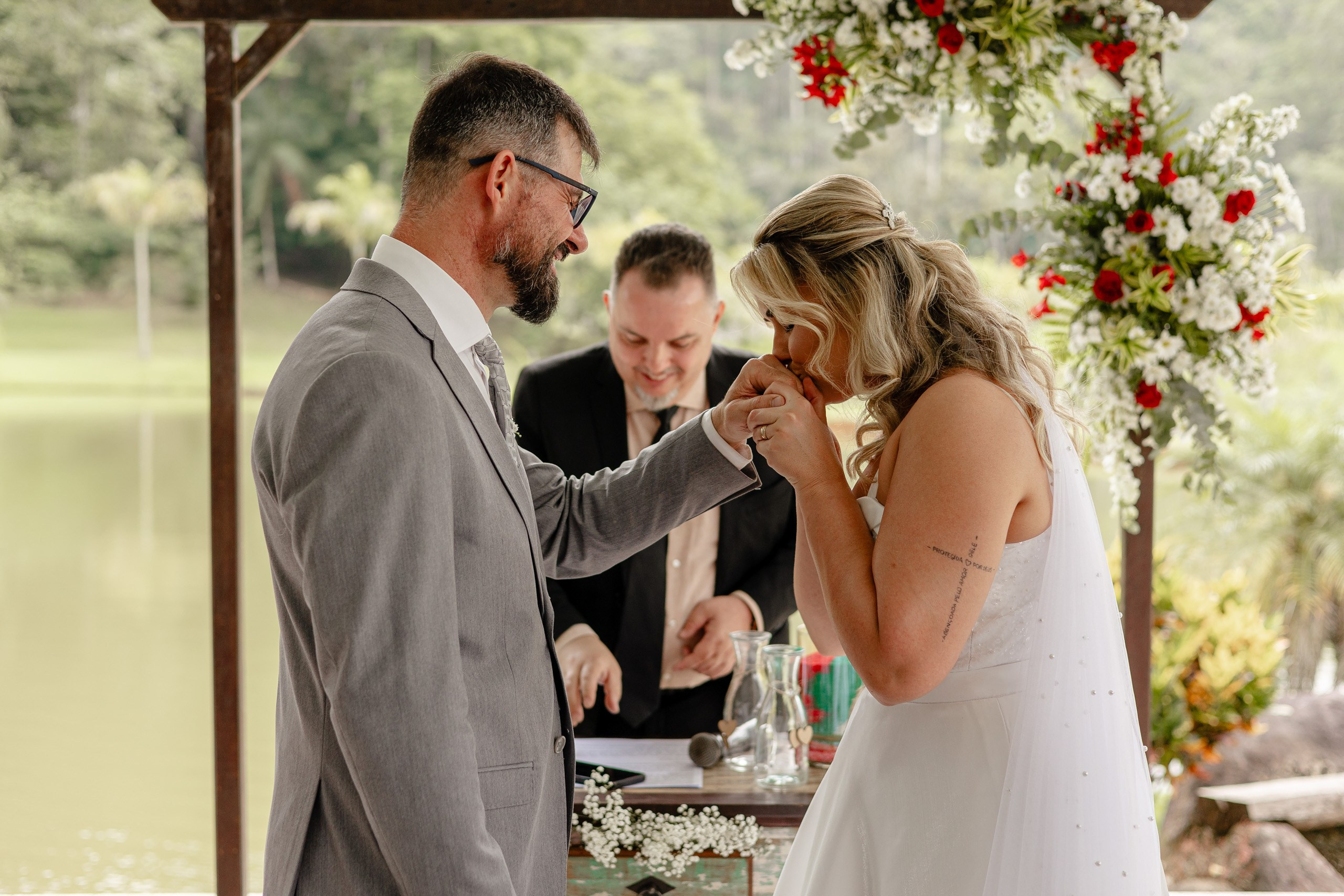 Fotógrafo de casamento em Santa Catarina; noivo beijando a mão da noiva emocionada diante do altar com arranjo floral durante cerimônia ao ar livre.