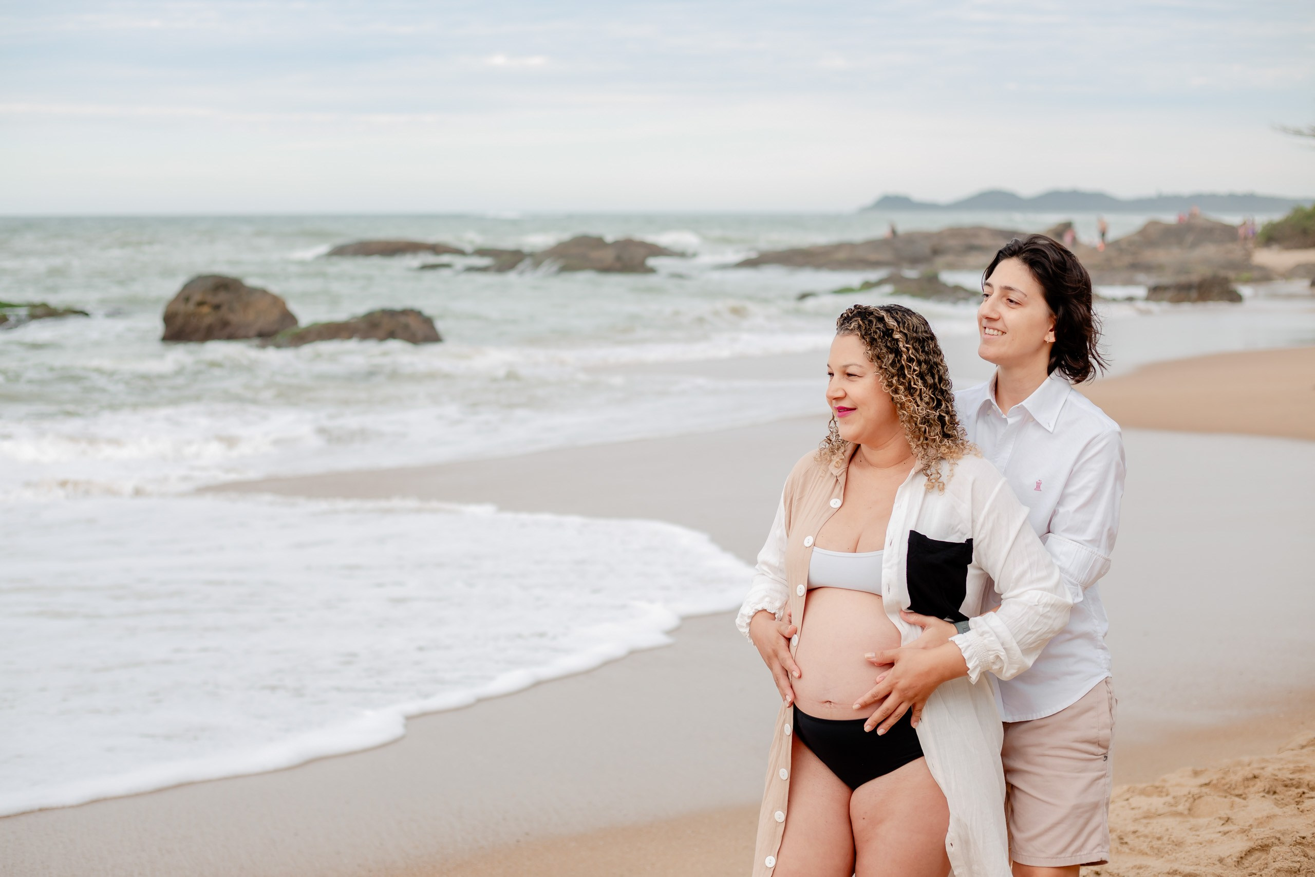 Fotógrafo ensaio gestante em Penha; casal abraçado contemplando o mar na praia com as mãos na barriga, em um registro leve e afetuoso à beira-mar.