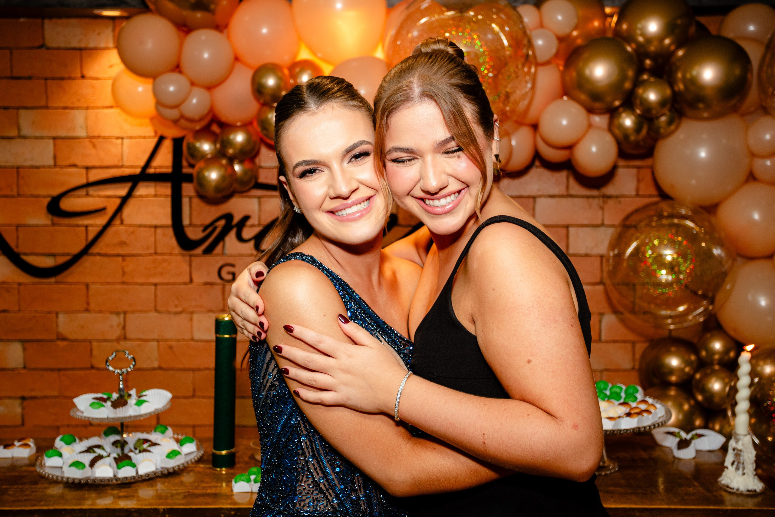 Fotógrafo de formatura em Balneário Camboriú; a formanda e uma amiga celebram abraçadas e sorridentes diante da mesa de doces e balões dourados.