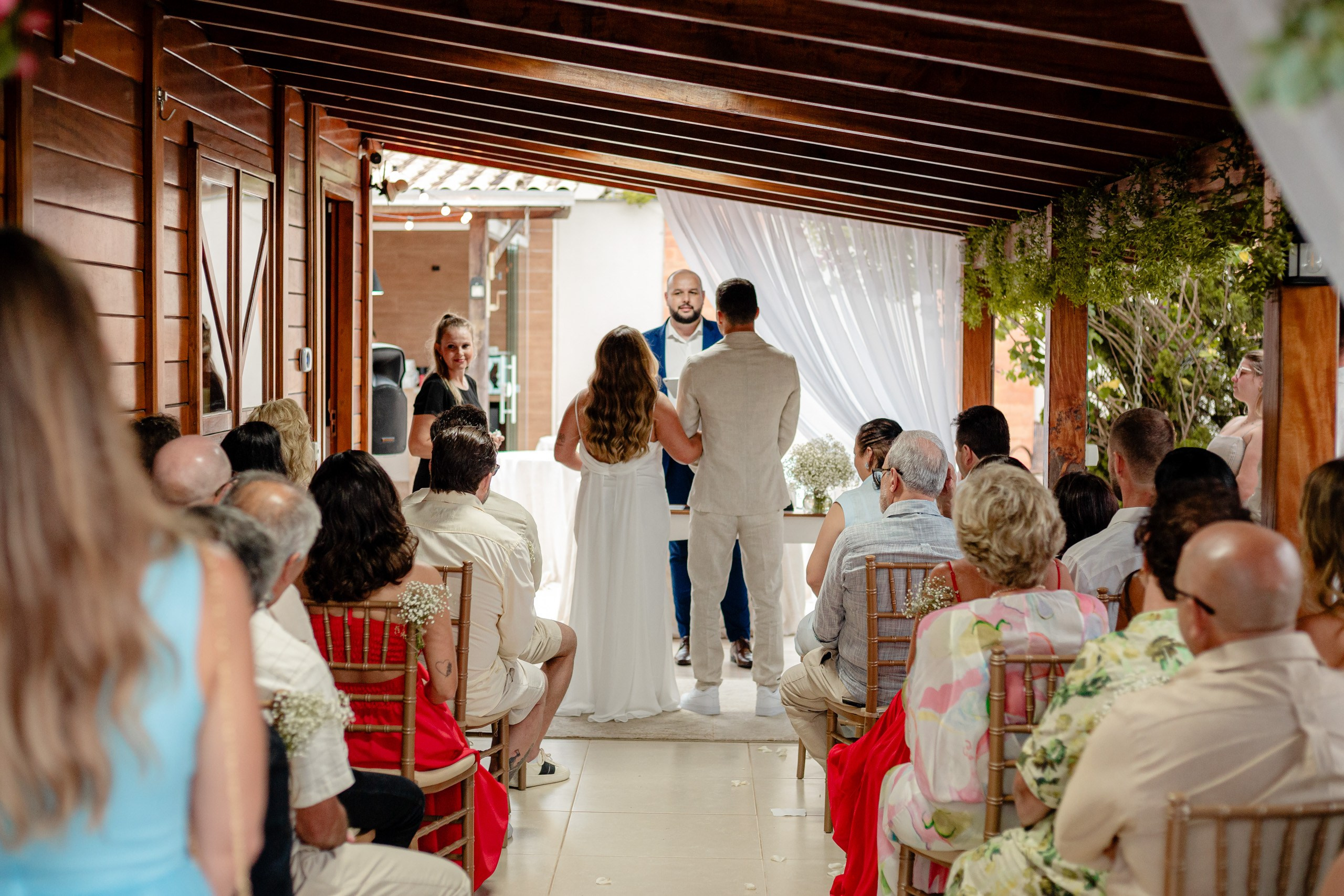 Fotógrafo de casamento em Bombinhas; visão geral da cerimônia ao ar livre sob uma estrutura de madeira, com o casal no altar diante de convidados atentos.
