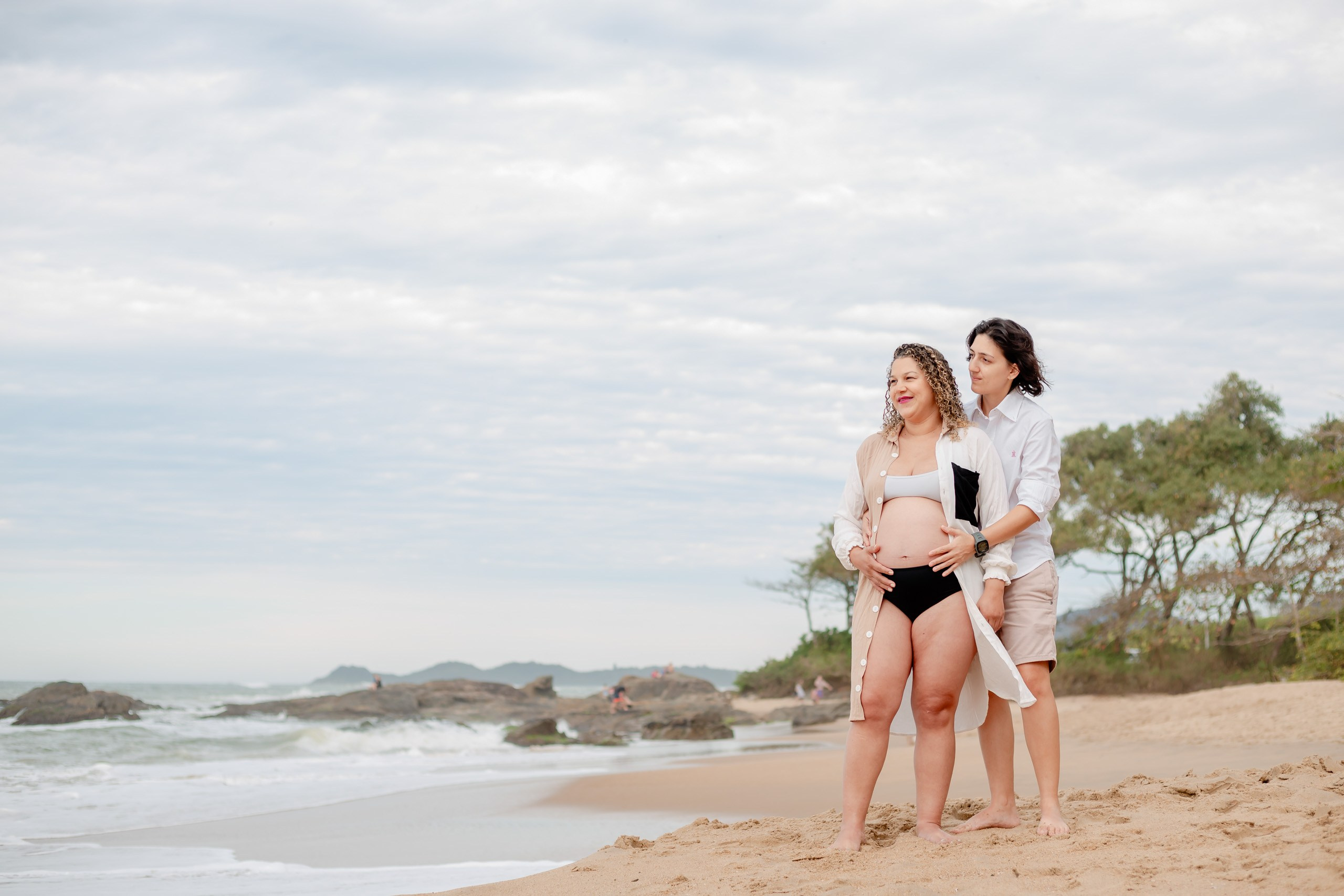 Fotógrafo ensaio gestante em Penha; plano geral do casal contemplando o mar e as formações rochosas na praia em um ensaio lifestyle litorâneo.