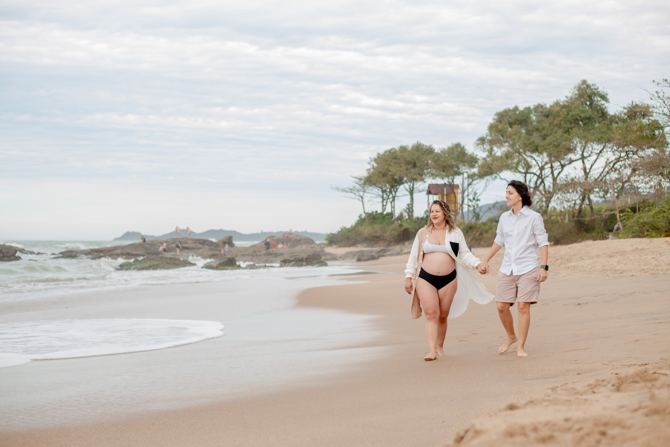 Fotógrafo ensaio gestante em Penha; casal caminhando sorridente de mãos dadas à beira-mar em um registro leve e espontâneo na praia.