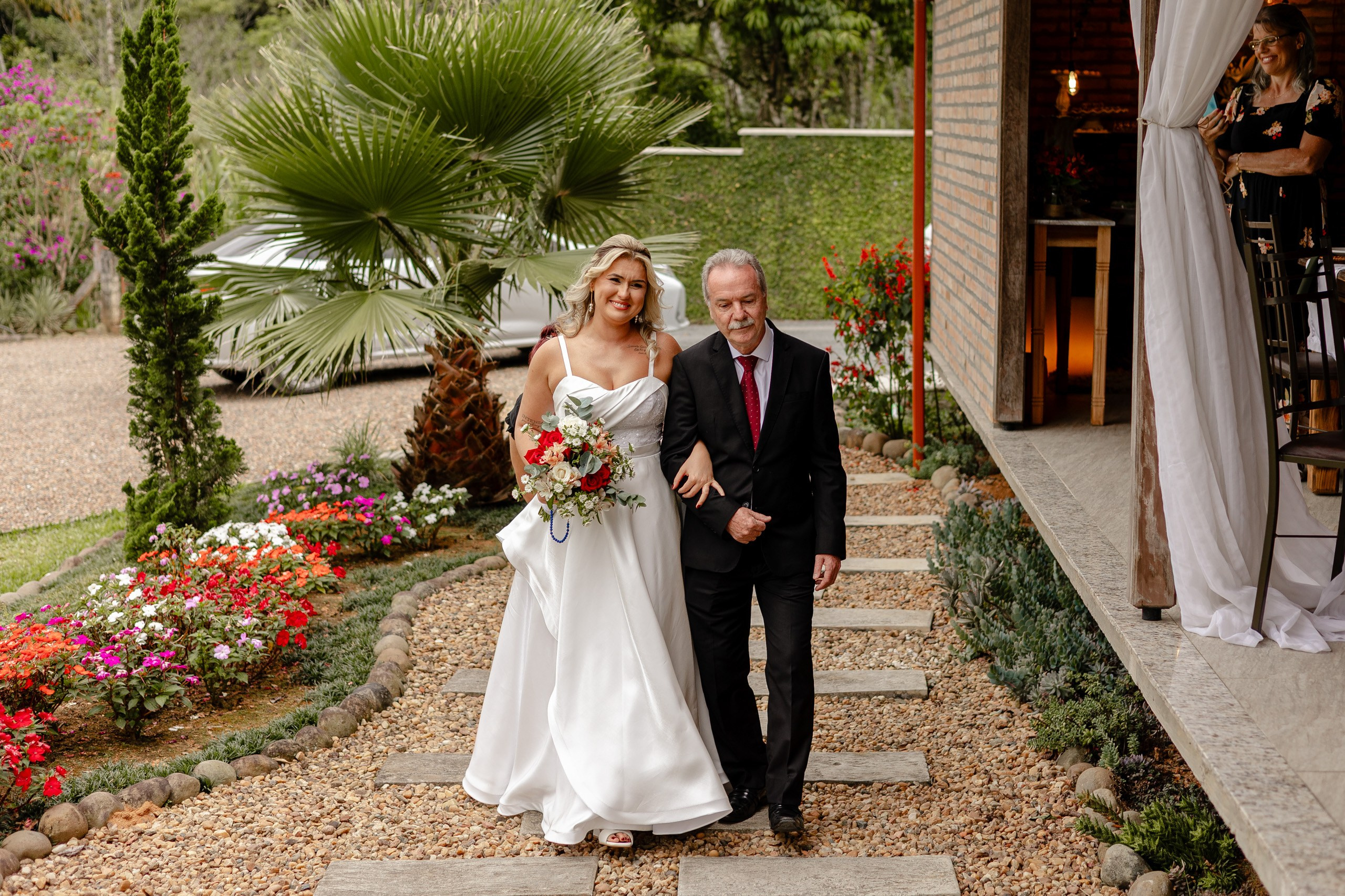 Fotógrafo de casamento em Santa Catarina; noiva de braço dado com familiar caminhando pelo caminho de pedras em direção à cerimônia ao ar livre.