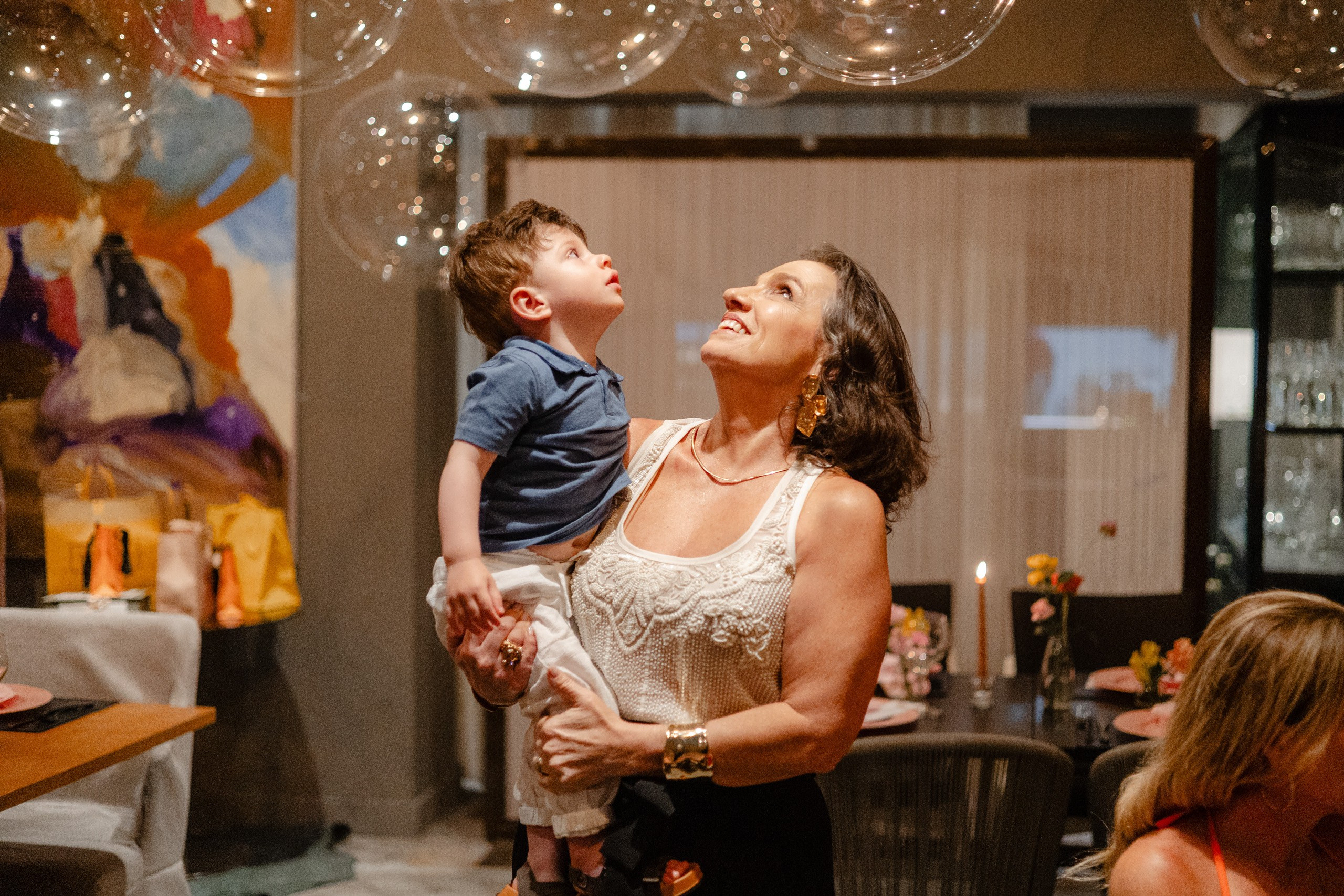 Fotógrafo de festa de formatura em Balneário Camboriú; mulher com criança no colo olhando para cima sob balões transparentes decorativos.