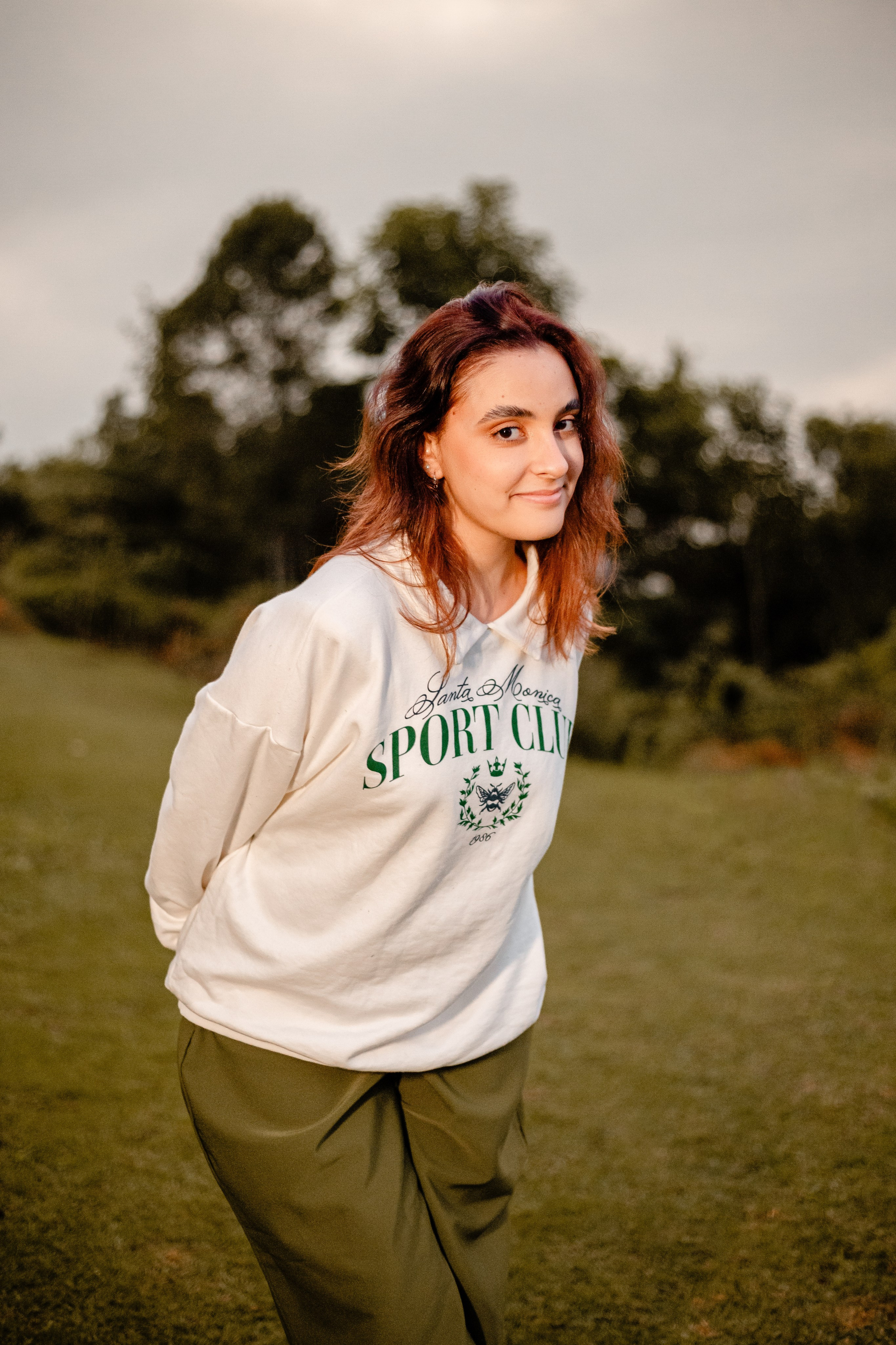 Fotógrafo de ensaio em Balneário Camboriú; retrato ao ar livre de jovem com cabelo acobreado, sweat branca com inscrição verde e calças verde-oliva, posando num ambiente natural e campestre.