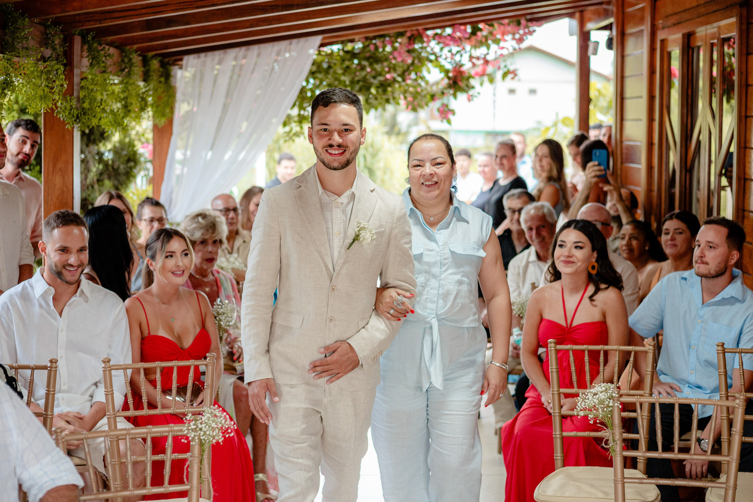 Fotógrafo de casamento em Bombinhas; o noivo caminha sorridente de braço dado durante a cerimônia ao ar livre, cercado por convidados atentos.